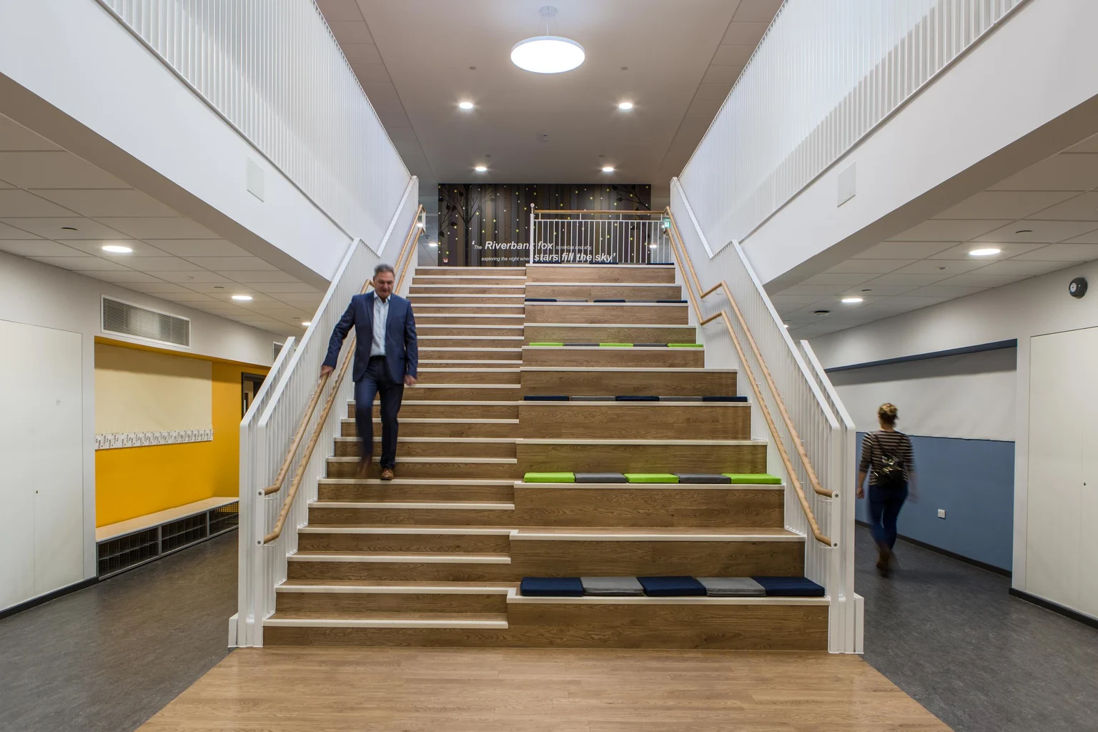 Un intérieur spacieux et moderne avec un large escalier intégrant des coins salon, rehaussé par des coussins colorés. L’espace est bien éclairé par des plafonniers et des puits de lumière, entouré de murs blancs immaculés et de détails jaunes éclatants.