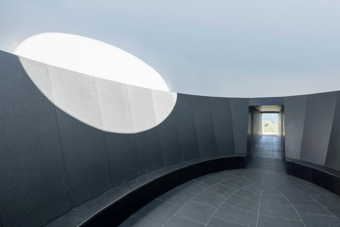 Ein ovaler Raum mit grauen Wänden und einem großen, runden Oberlicht, das Tageslicht hineinlässt. Am Ende des Raums führt ein Durchgang zu einer Aussicht auf eine grüne Landschaft.