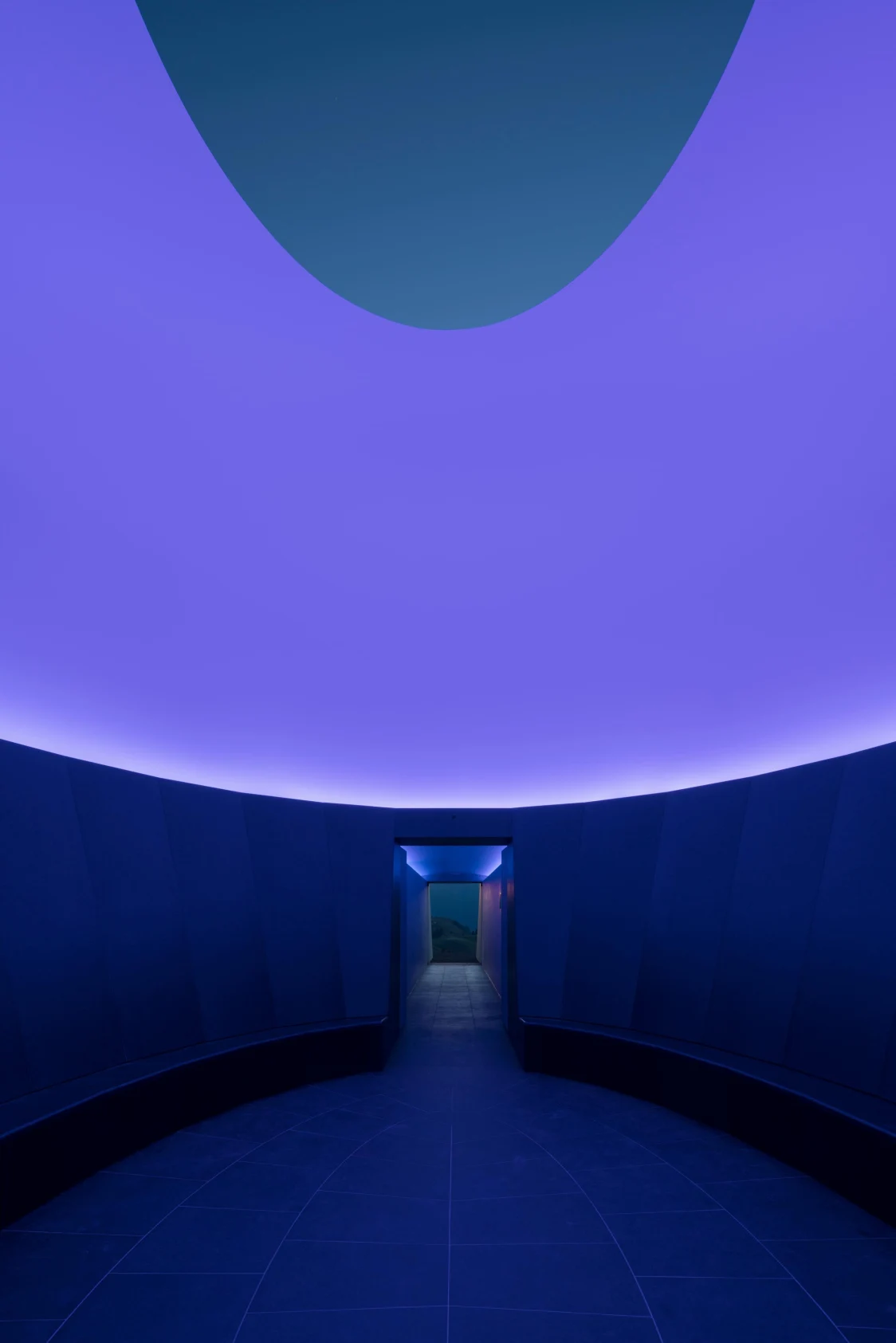 Ein dunkler, ovaler Raum mit leuchtenden, violetten Lichtakzenten an der Decke führt durch einen schmalen Durchgang zu einer grünen Landschaft. Die futuristische Beleuchtung schafft eine atmosphärische und beruhigende Stimmung.