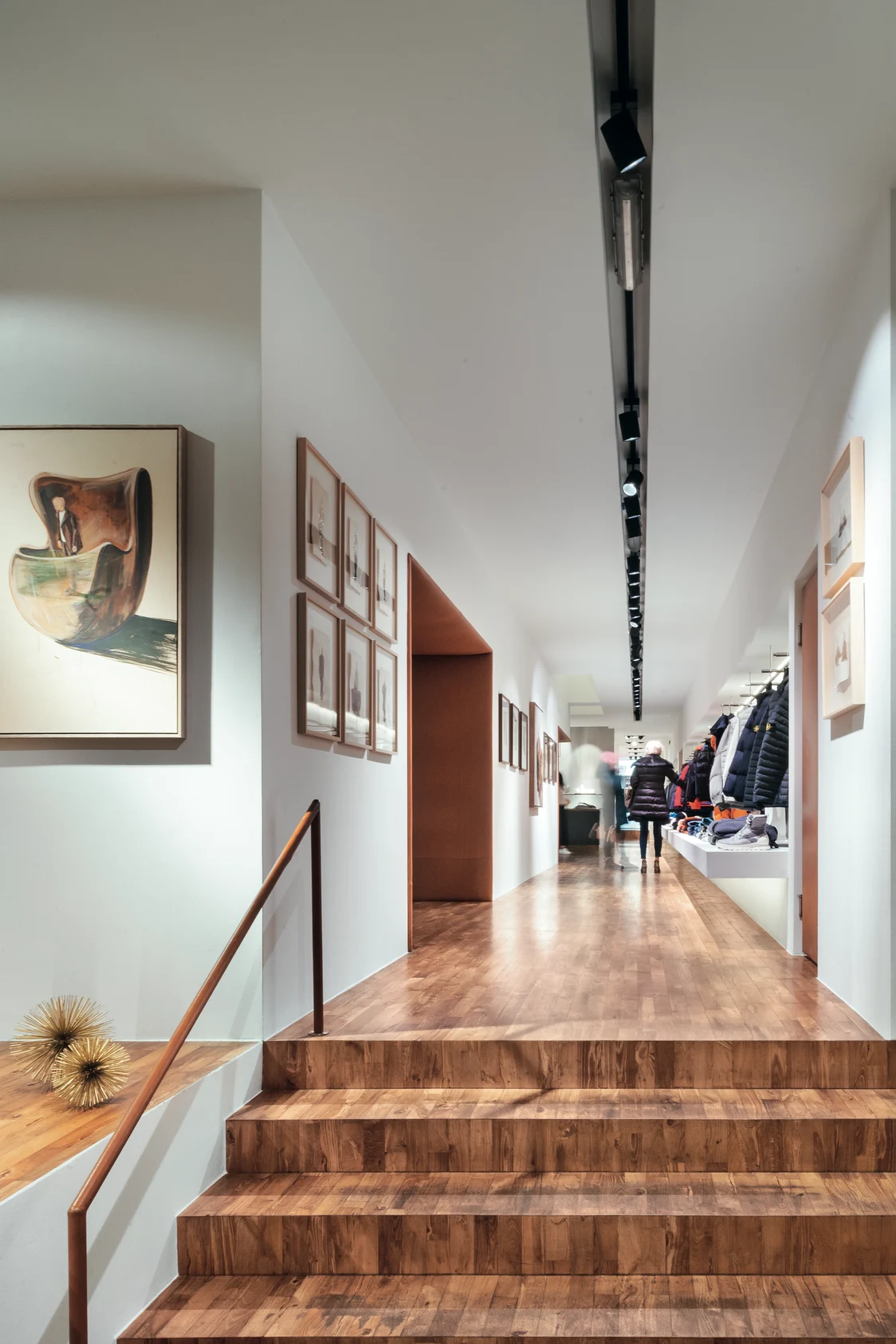 TRACKS-Beleuchtung erhellt einen langen Flur mit Holzboden in einem modernen Laden, flankiert von Bildern und rechts ausgestellten Jacken.