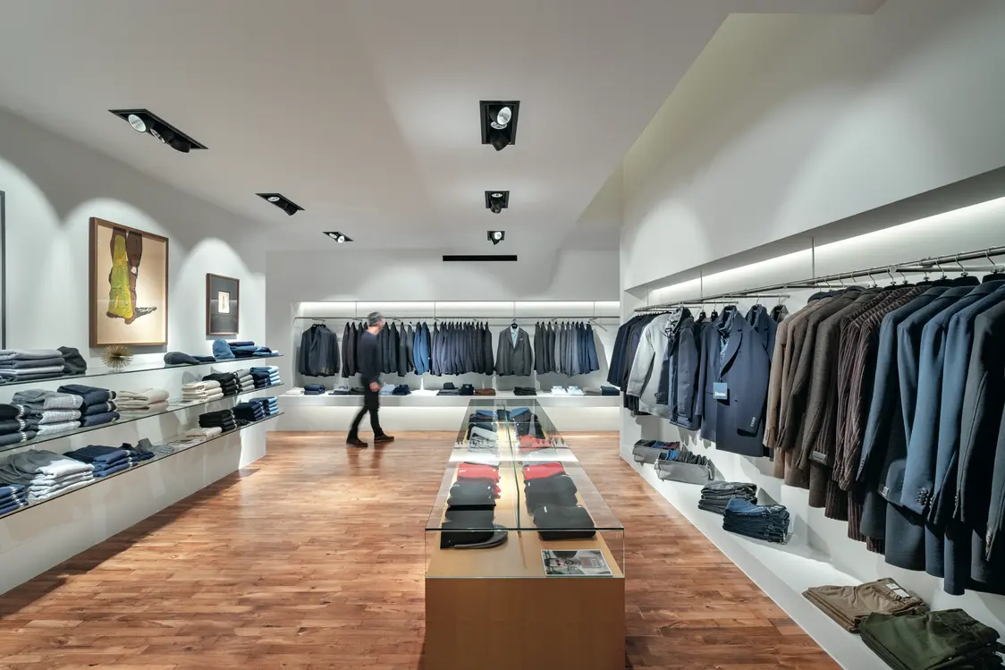 Un moderno negozio da uomo con pavimenti in legno, illuminazione elegante e abbigliamento di alta qualità ben esposti su appendiabiti. In primo piano c'è un tavolo con abiti piegati e accessori.