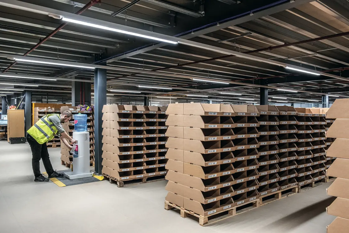Ein Mitarbeiter in Warnweste bedient eine Maschine in einem großen, gut beleuchteten Lager mit vielen gestapelten Kartons auf Paletten. Die Umgebung wirkt ordentlich und organisiert.