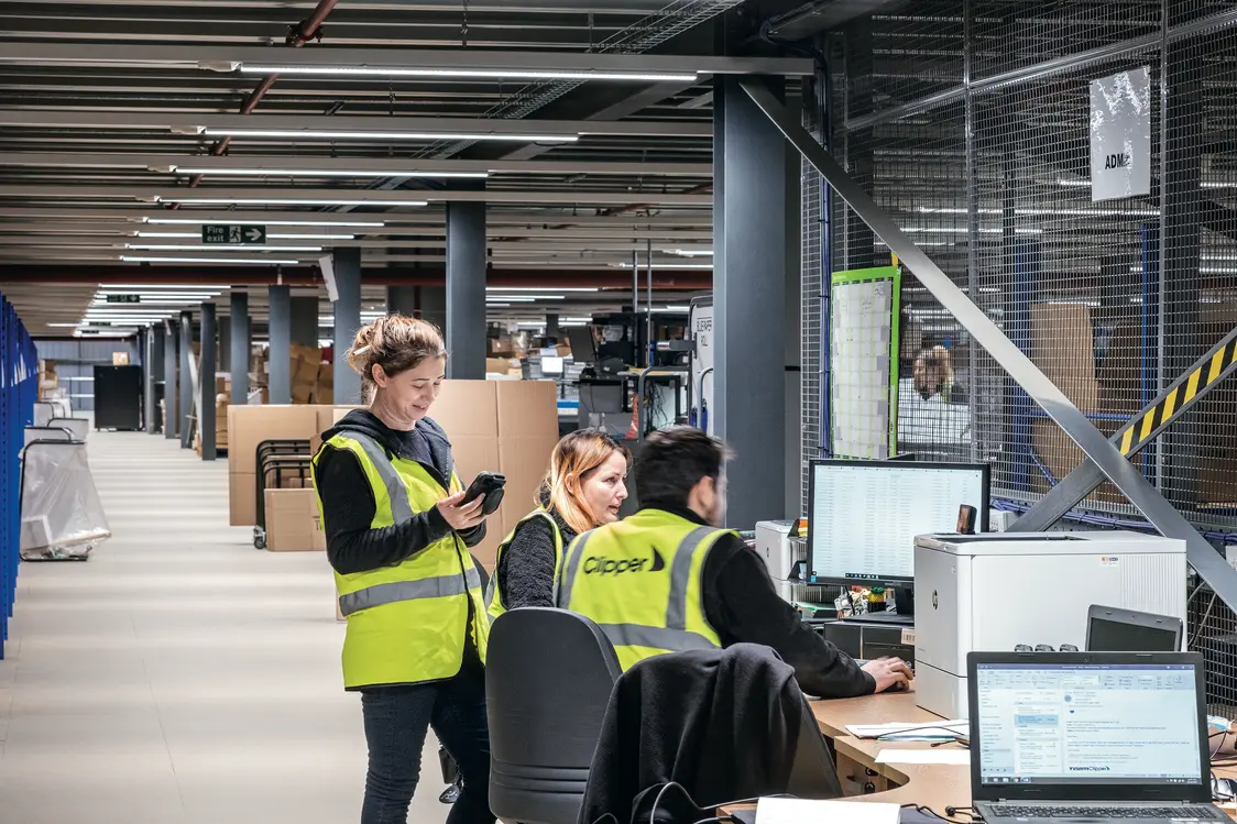 Drei Personen in gelben Warnwesten arbeiten in einem modernen Lagerbüro an Computern und mit mobilen Geräten. Im Hintergrund sind Regale und Lagerflächen zu sehen, die eine organisierte Arbeitsumgebung zeigen.
