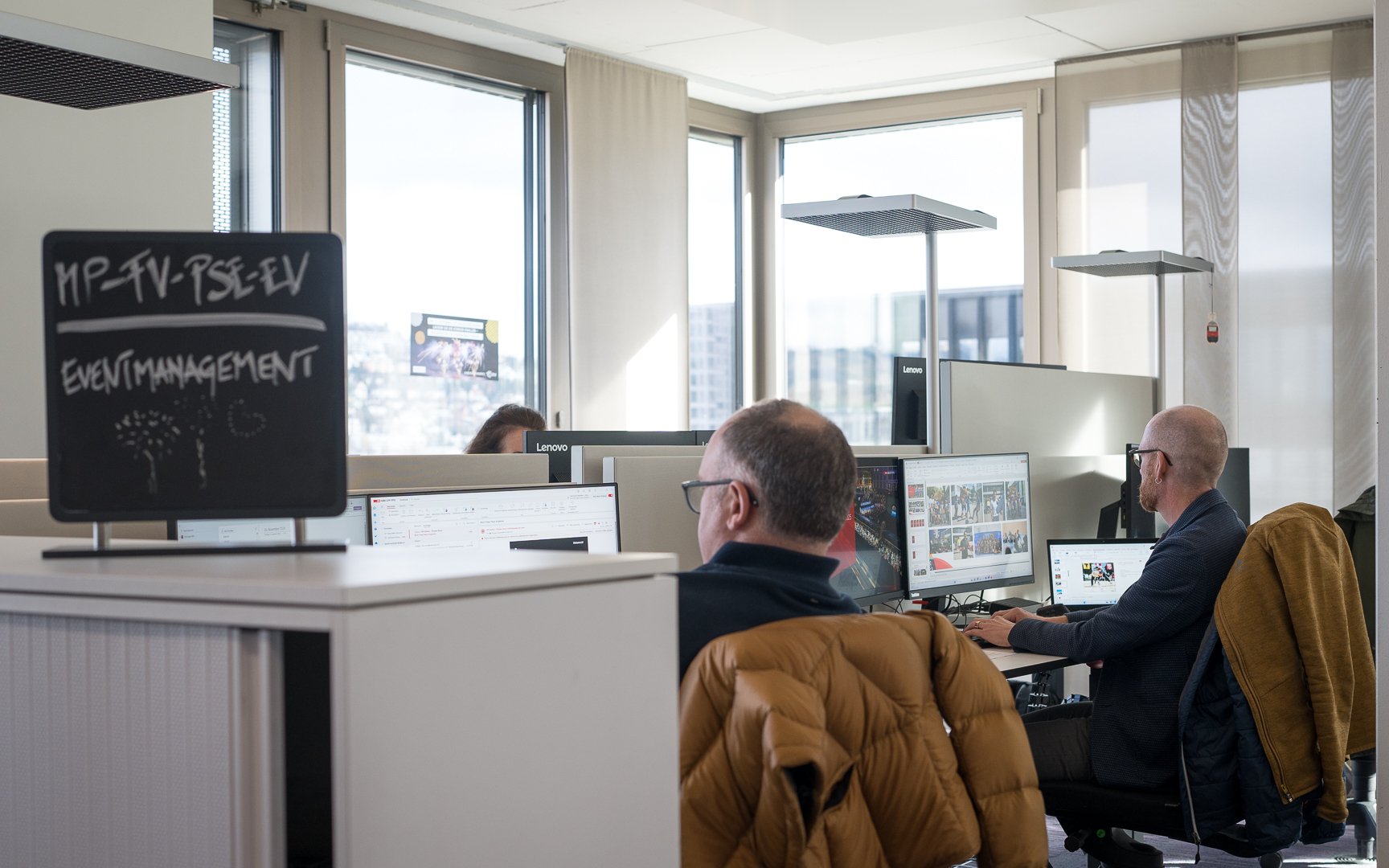 Drei Personen sitzen vor je einem Computer. Die Sonne scheint ins Büro