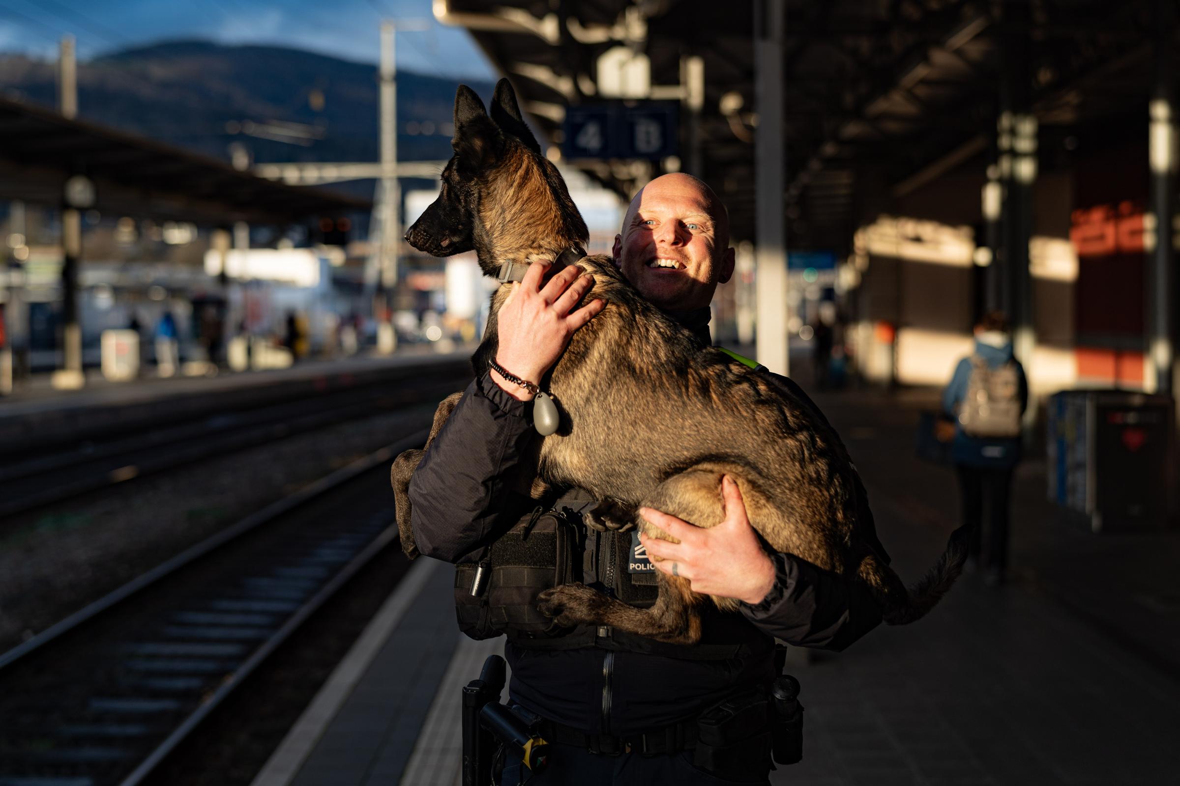 Der Polizist steht auf dem Perron am Bahnhof und hebt den Schäferhund in den Armen.