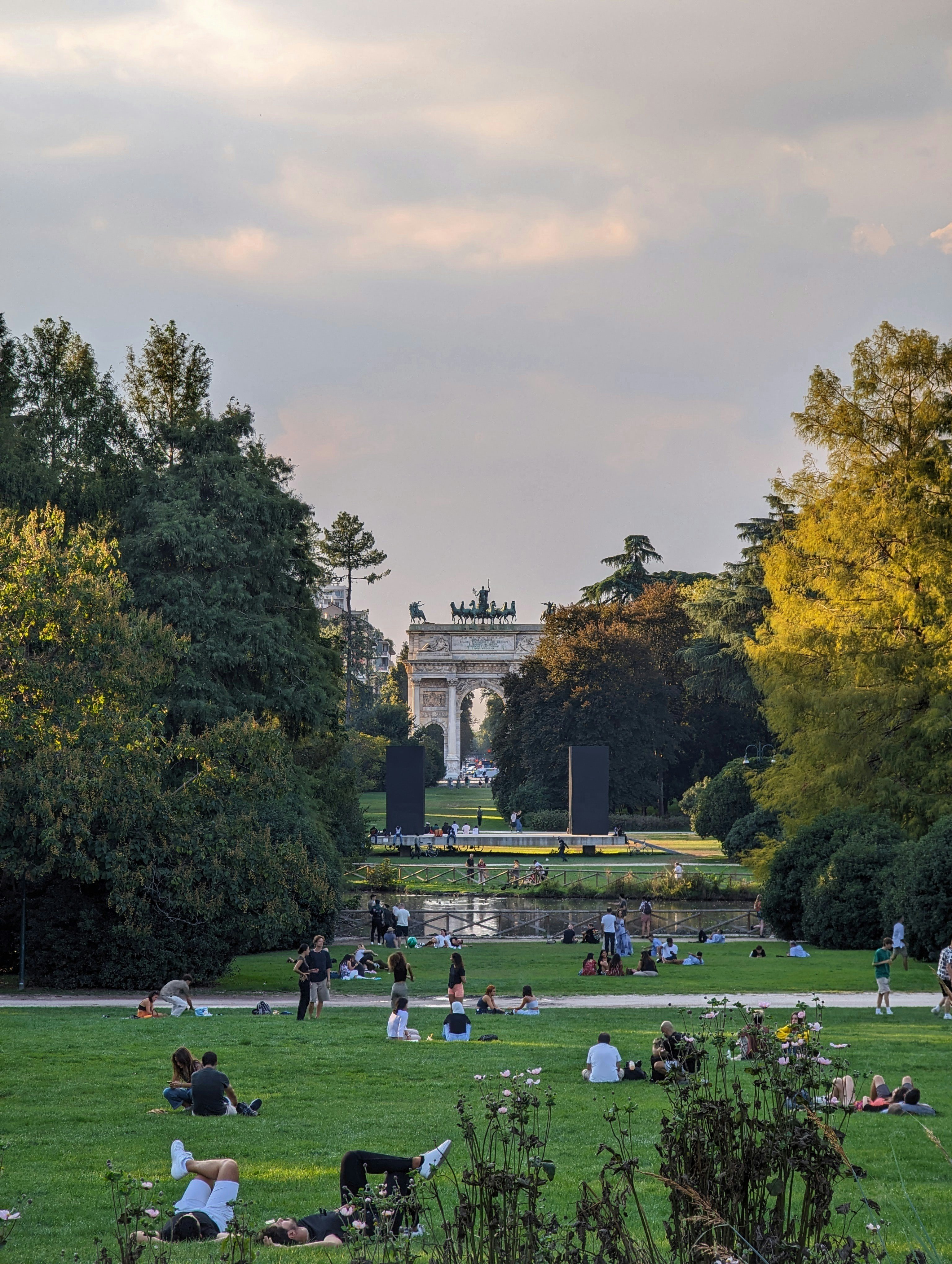 Personen liegen und sitzen auf einem Rasen in einem Park. Im Hintergrund ist ein bogenähnliches Bauwerk zu sehen.