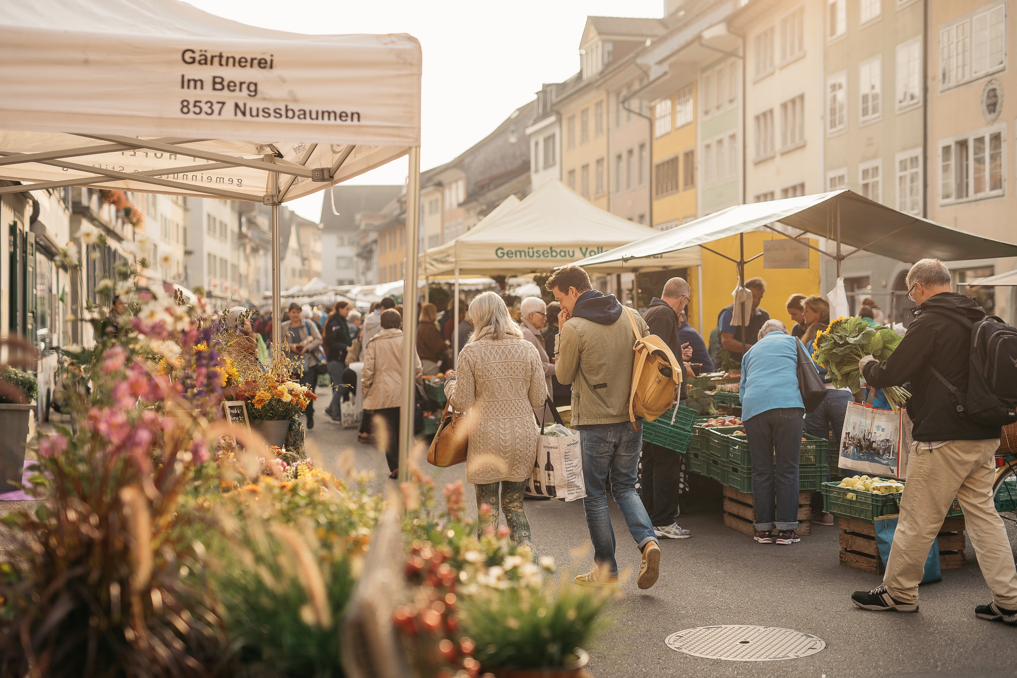 Menschen durchqueren die Marktstände mit Blumen und Gemüse vor den Fassaden der Altstadthäuser.