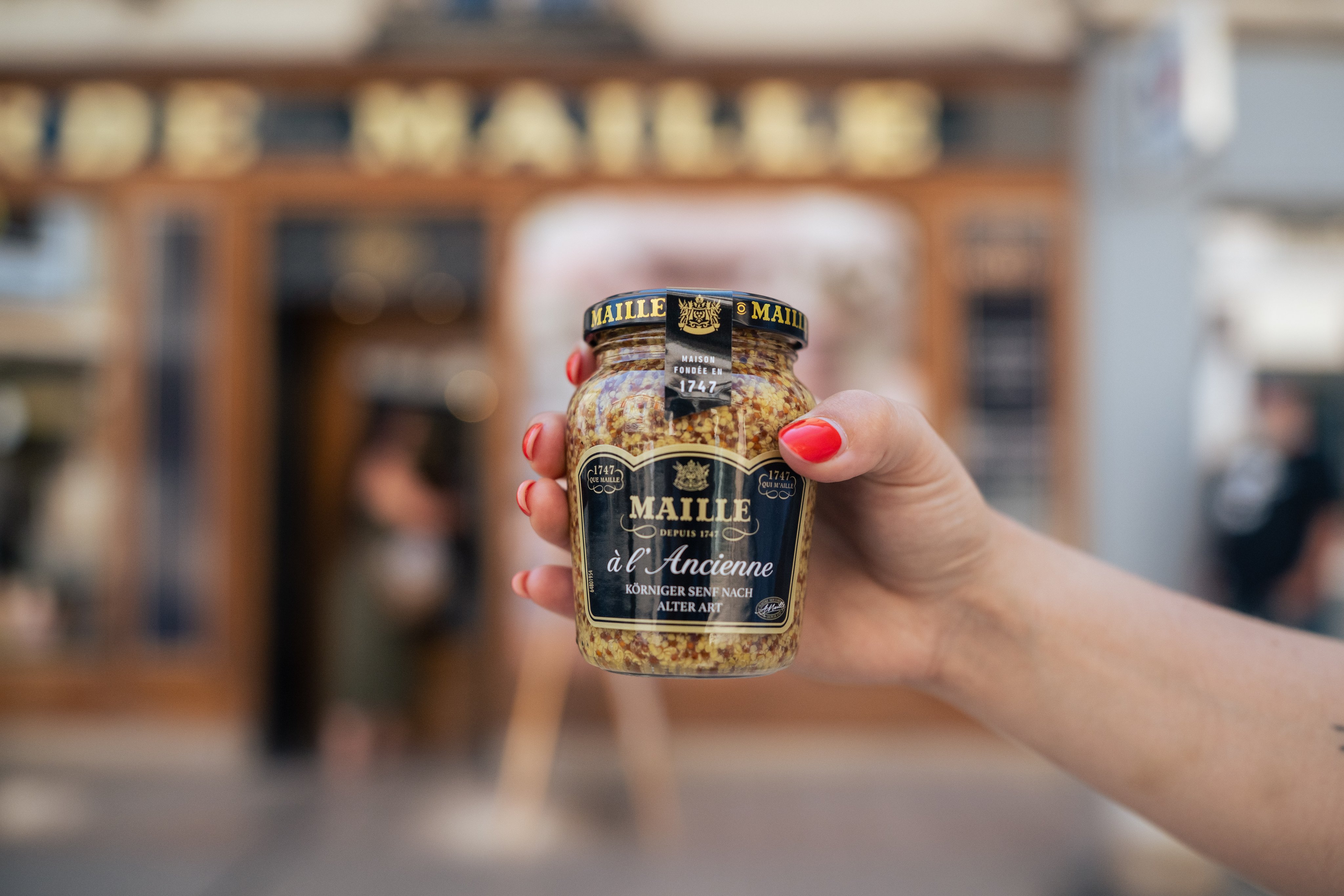 Eine Hand hält ein Glas mit dem originalen körnigen Maille-Senf in die Luft.