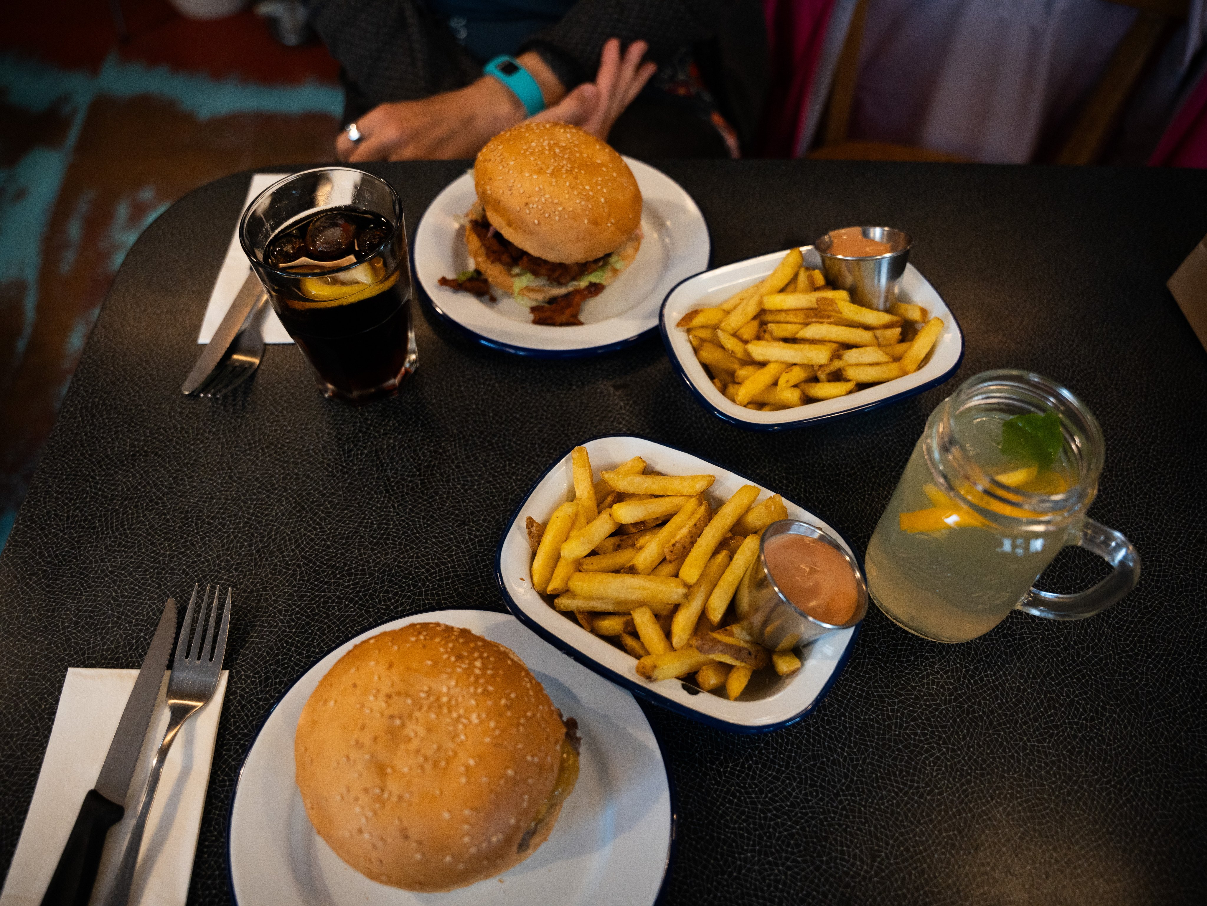 Teller und Schüsseln mit Hamburgern, Salat und Pommes Frites stehen auf einem Tisch, ebenso zwei Gläser mit Getränken.