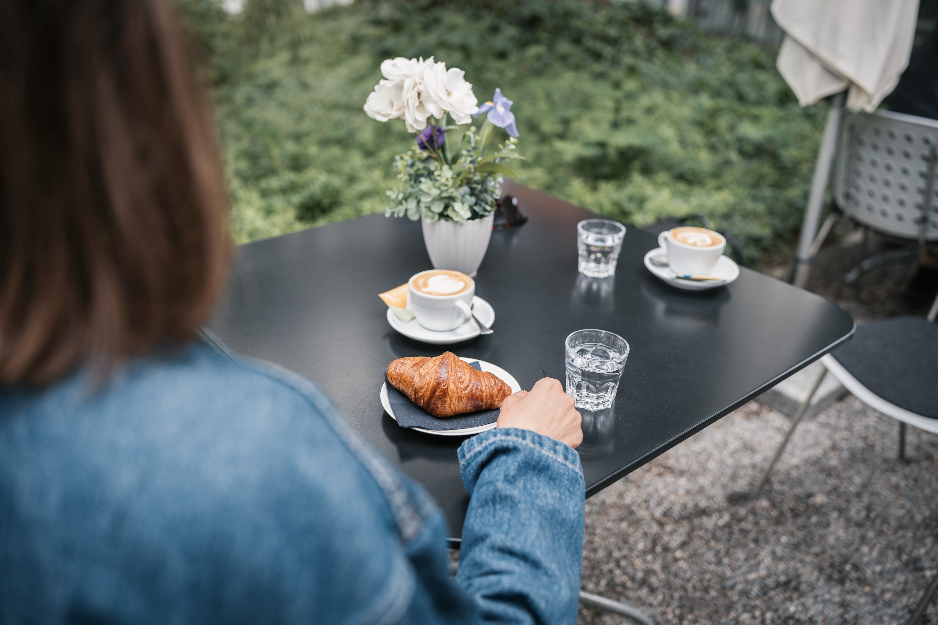 Frau sitzt an einem Gartentisch und geniesst Cappuccino und Buttergipfel.
