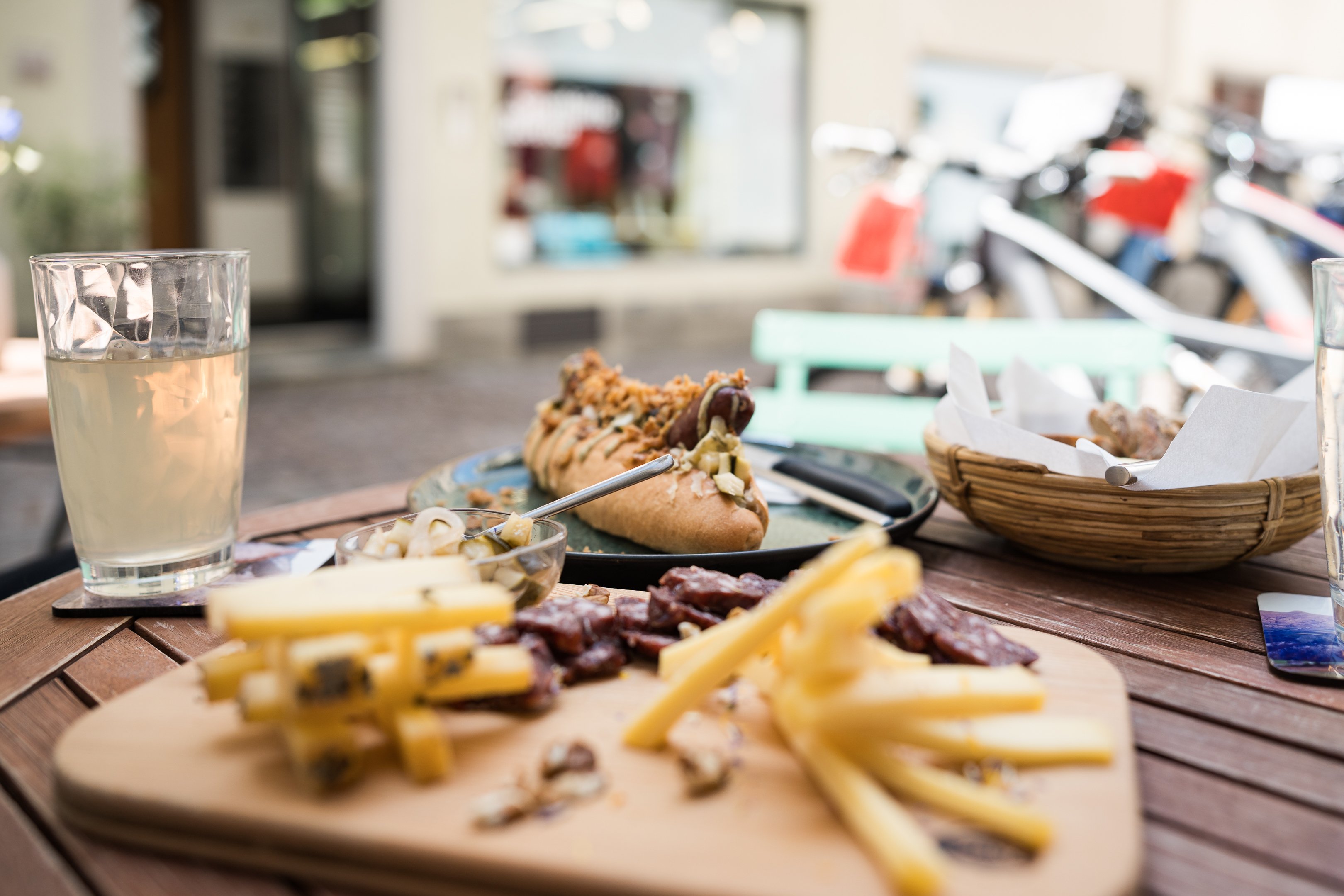 Gedeckter Tisch im Freien mit einem Hotdog, einer Wurst-Käse-Platte und einem Glas Eistee.