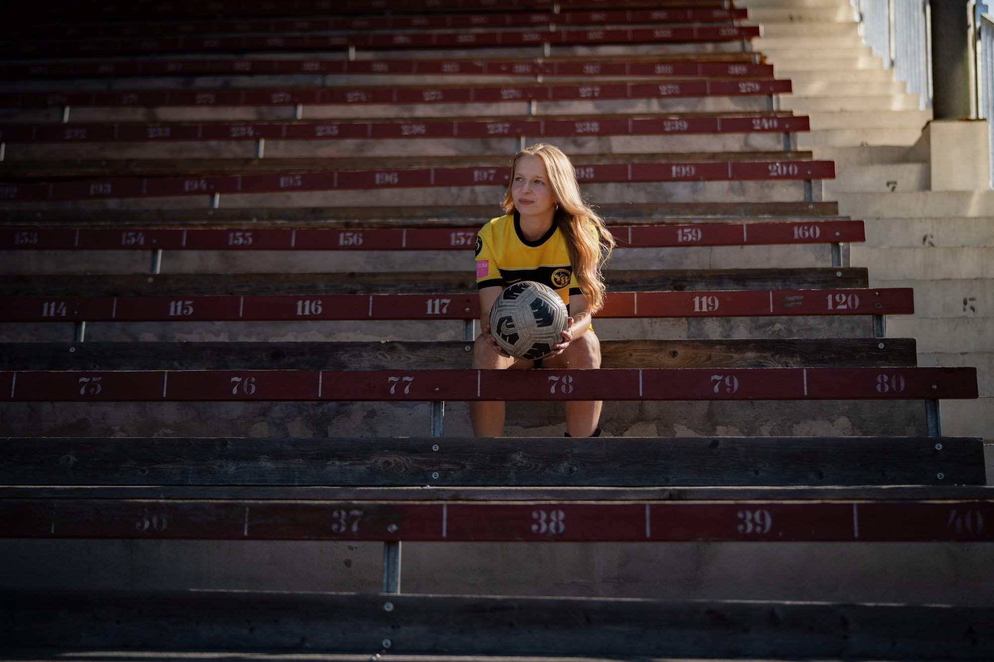 Une femme au regard rêveur, en tenue de sport, est assise dans une tribune avec un ballon dans les mains.