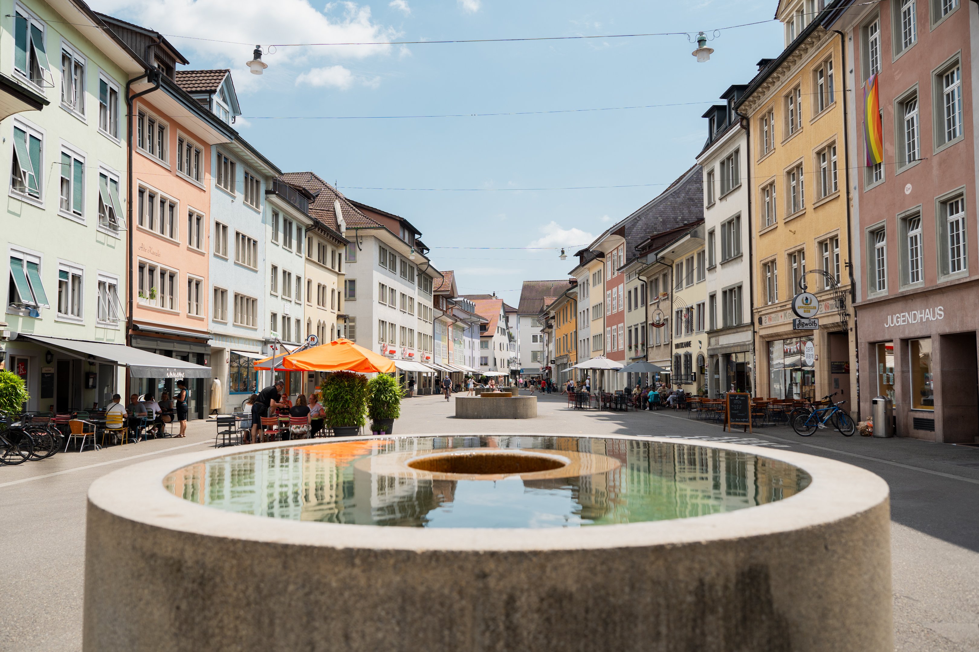 Runder Brunnen mit Loch in Mitte steht in Fussgängerzone. Gesäumt von bunten Häuserfassaden.