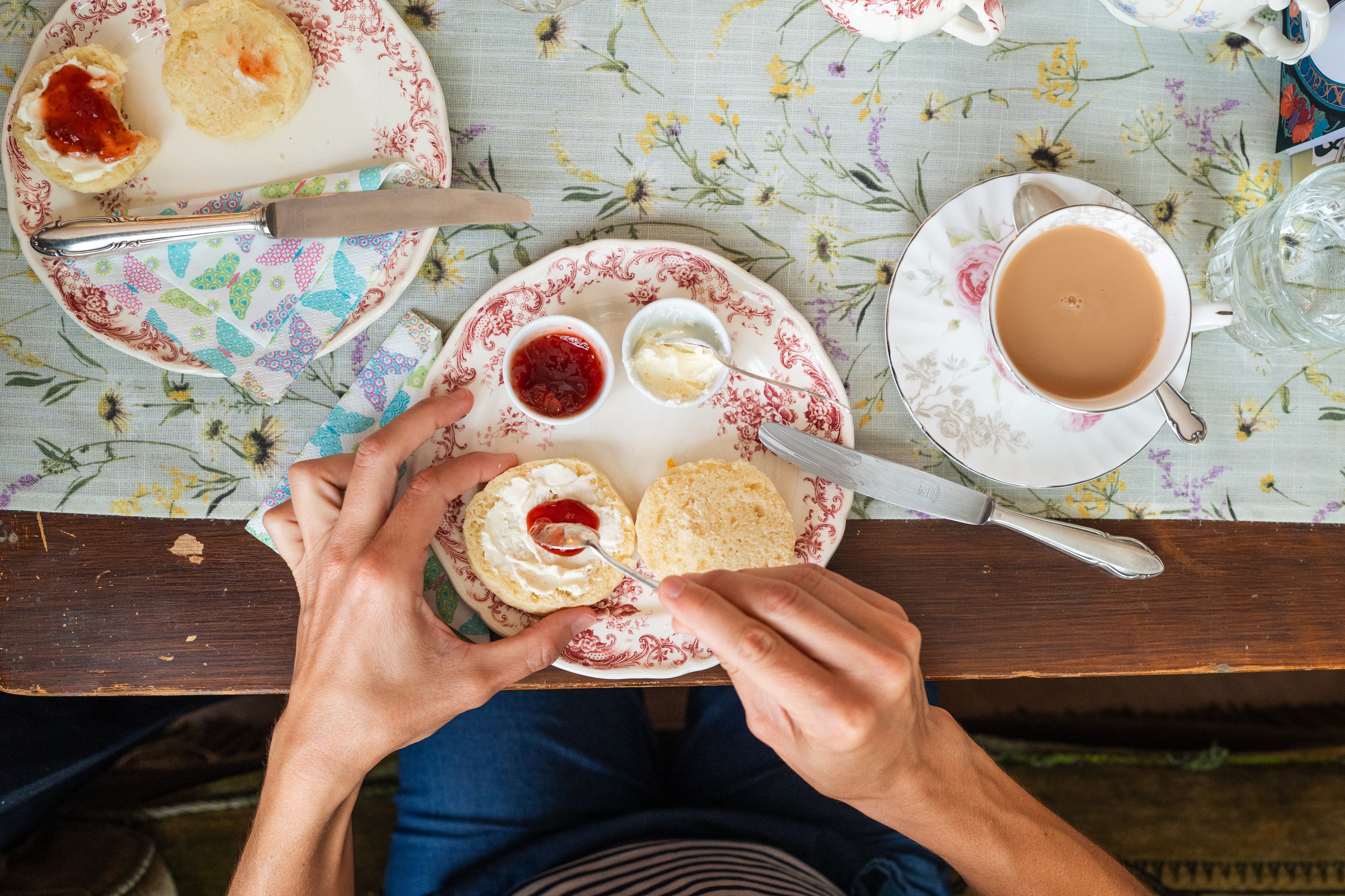 Vogelperspektive auf Porzellanteller mit Scones und Tasse Tee. Person bestreicht Scone mit Konfitüre.
