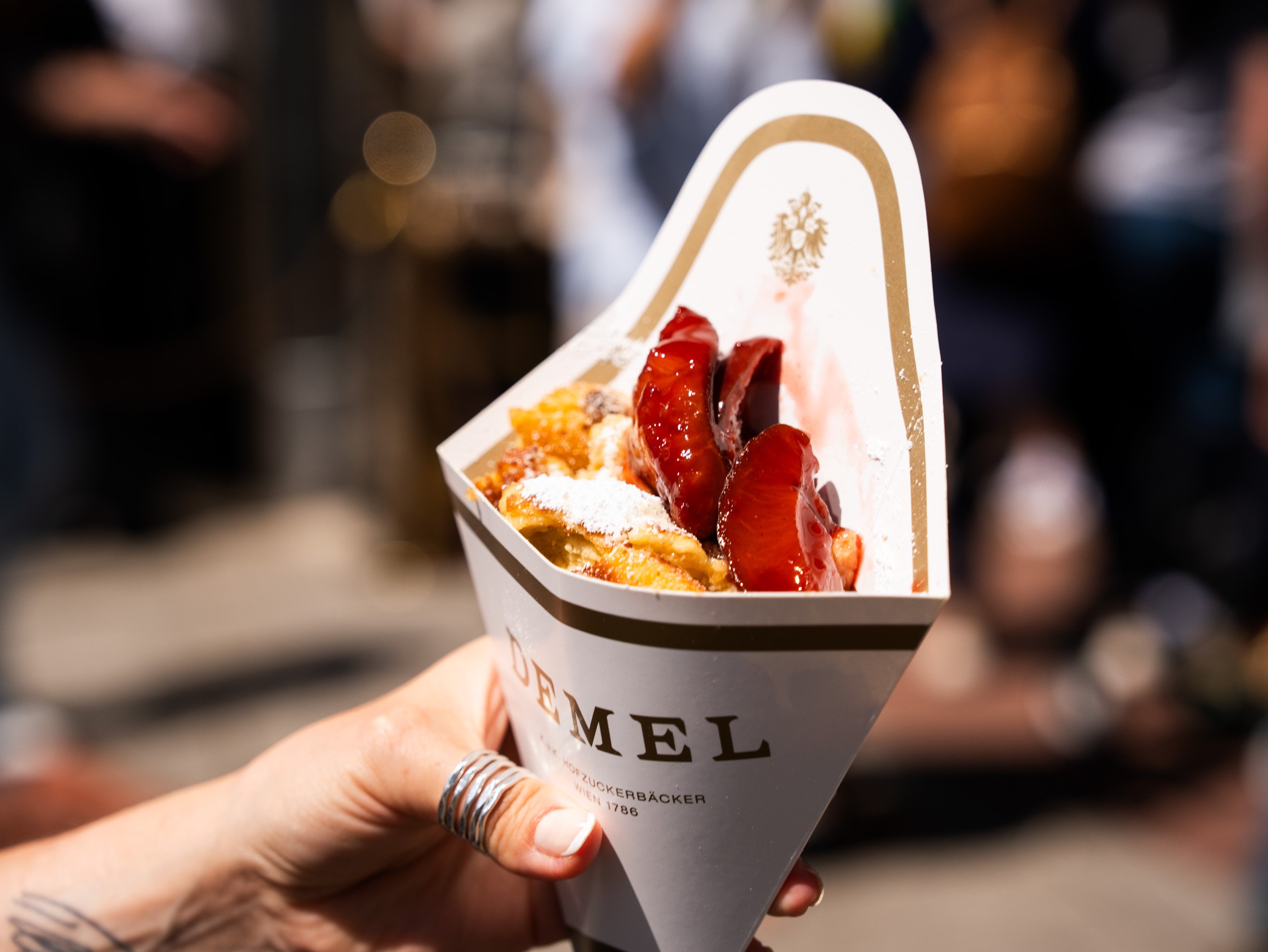 Une main tient un cornet en carton de l’ancienne pâtisserie de la cour viennoise Demel, contenant un Kaiserschmarren garni de compote de quetsches et de sucre glace.