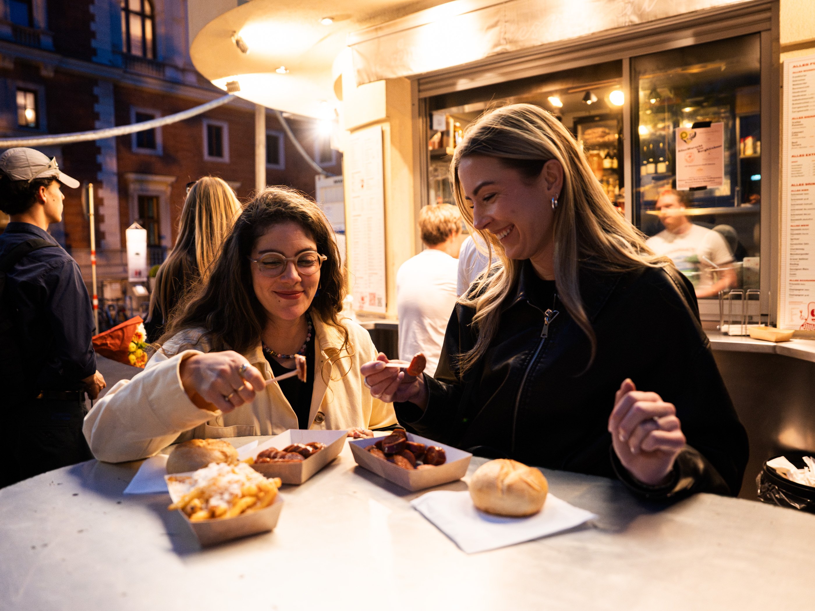 En soirée, deux femmes mangent des Käsekrainer avec des frites au Würstelstand viennois Alles Wurscht.