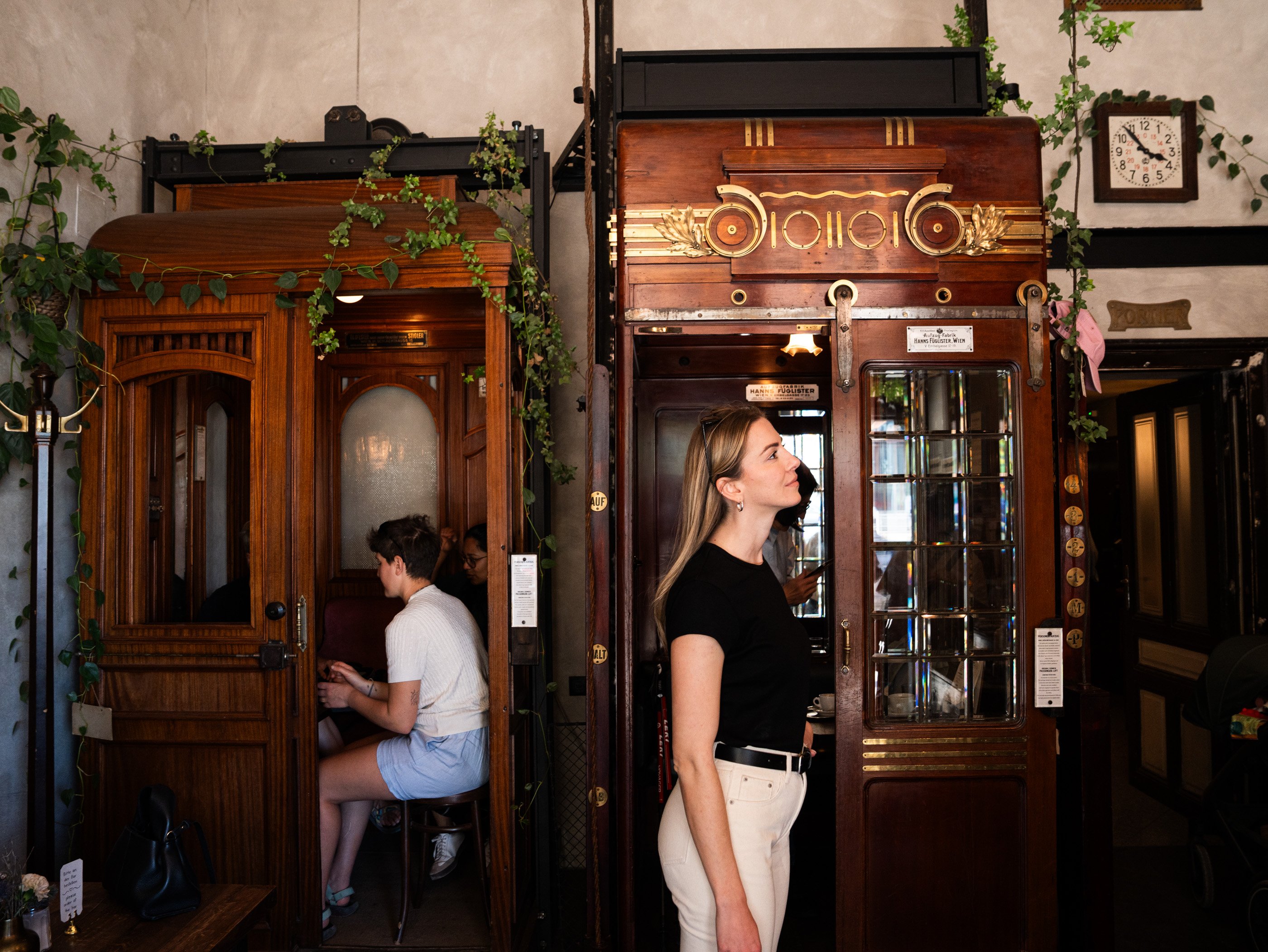 Une femme est debout à côté de deux cabines d’ascenseur historiques de l’Aufzug Café de Vienne, dans lesquelles des clients boivent un café.