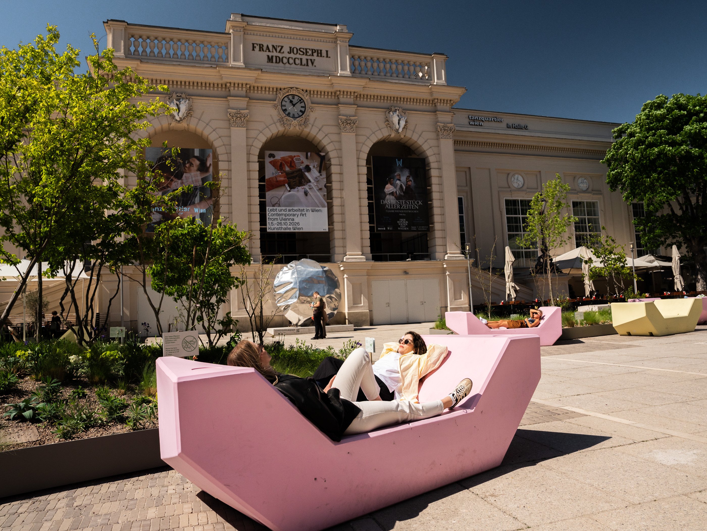 Par une journée ensoleillée, deux femmes sont allongées sur une sorte de banquette rose située devant un musée dans la cour intérieure du MuseumsQuartier, à Vienne.