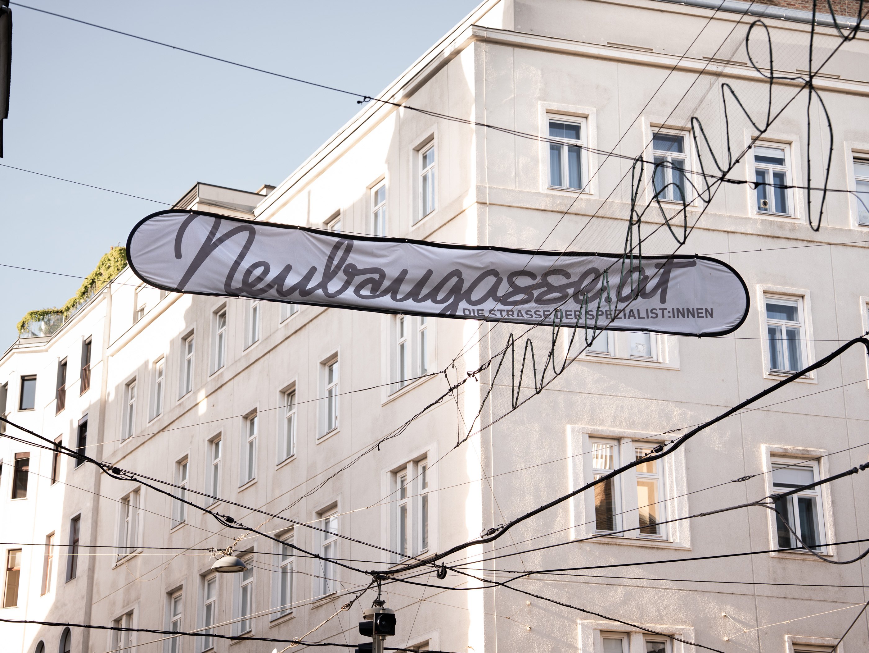 Une banderole sur laquelle est écrit «Neubaugasse.at» est suspendue au-dessus d’une rue viennoise, au milieu d’immeubles clairs et de lignes de contact de tram.
