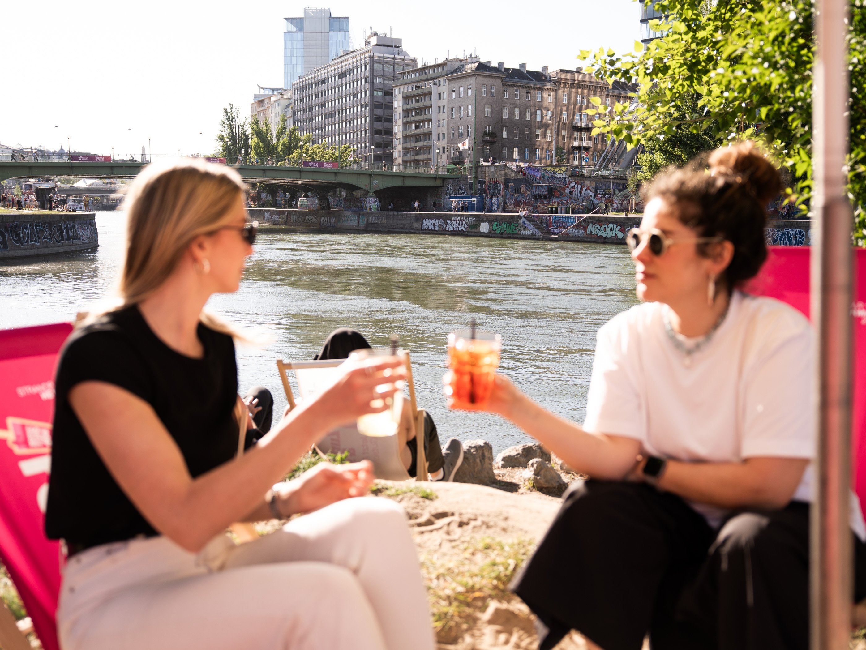 Deux femmes trinquent au Strandbar Herrmann, au bord du canal du Danube, à Vienne. On aperçoit un pont et des graffiti à l’arrière-plan.