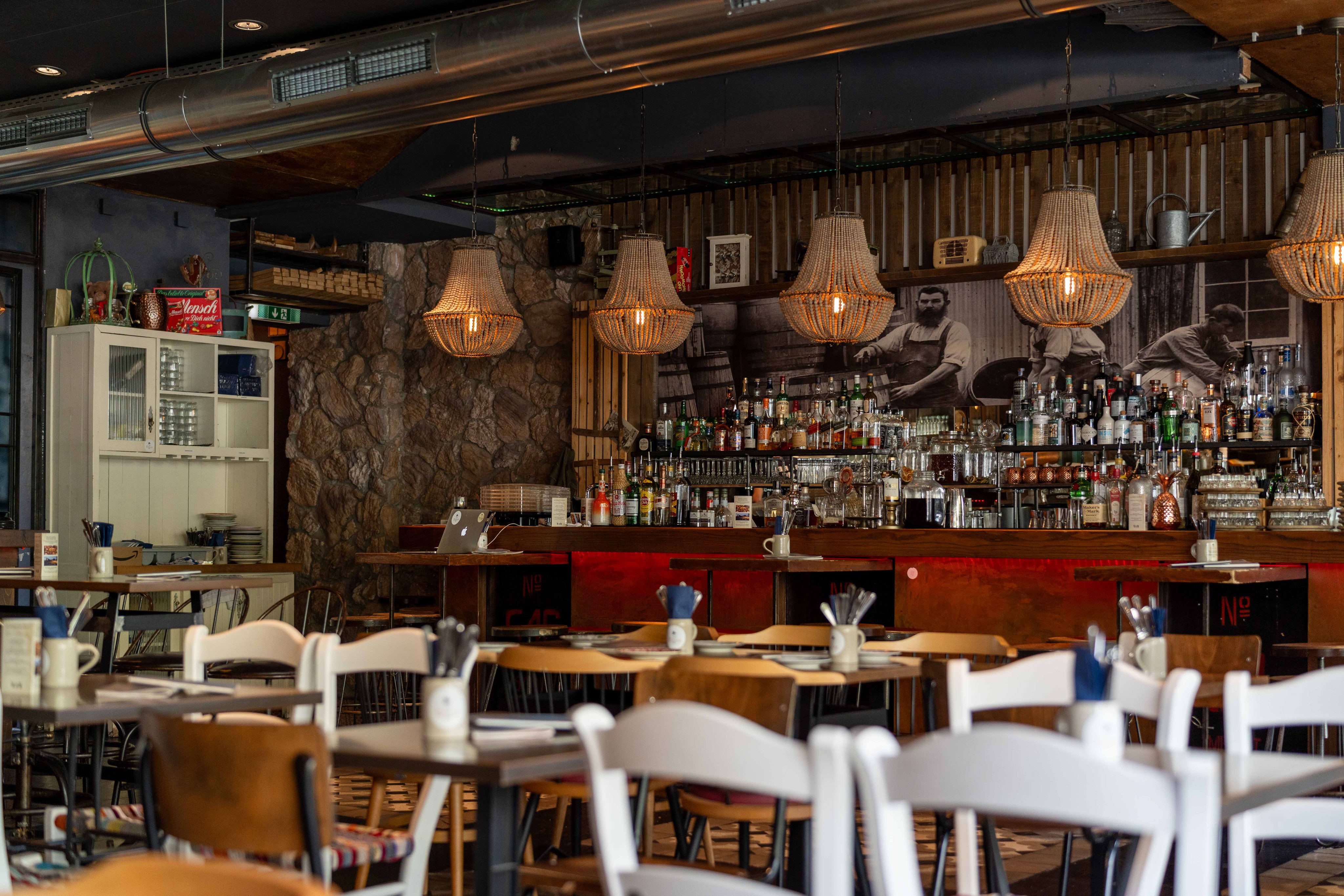 Die Innenseite des Restaurants: Eine Theke mit verschiedenen Flaschen und Gläsern auf den Regalen. Die Theke hat eine Steinwand auf einer Seite und eine Holzwand auf der anderen. Im Vordergrund sind Holztische und Stühle zu sehen. Die Decke hat freiliegende Rohre und hängende Lampen mit geflochtenen Schirmen.