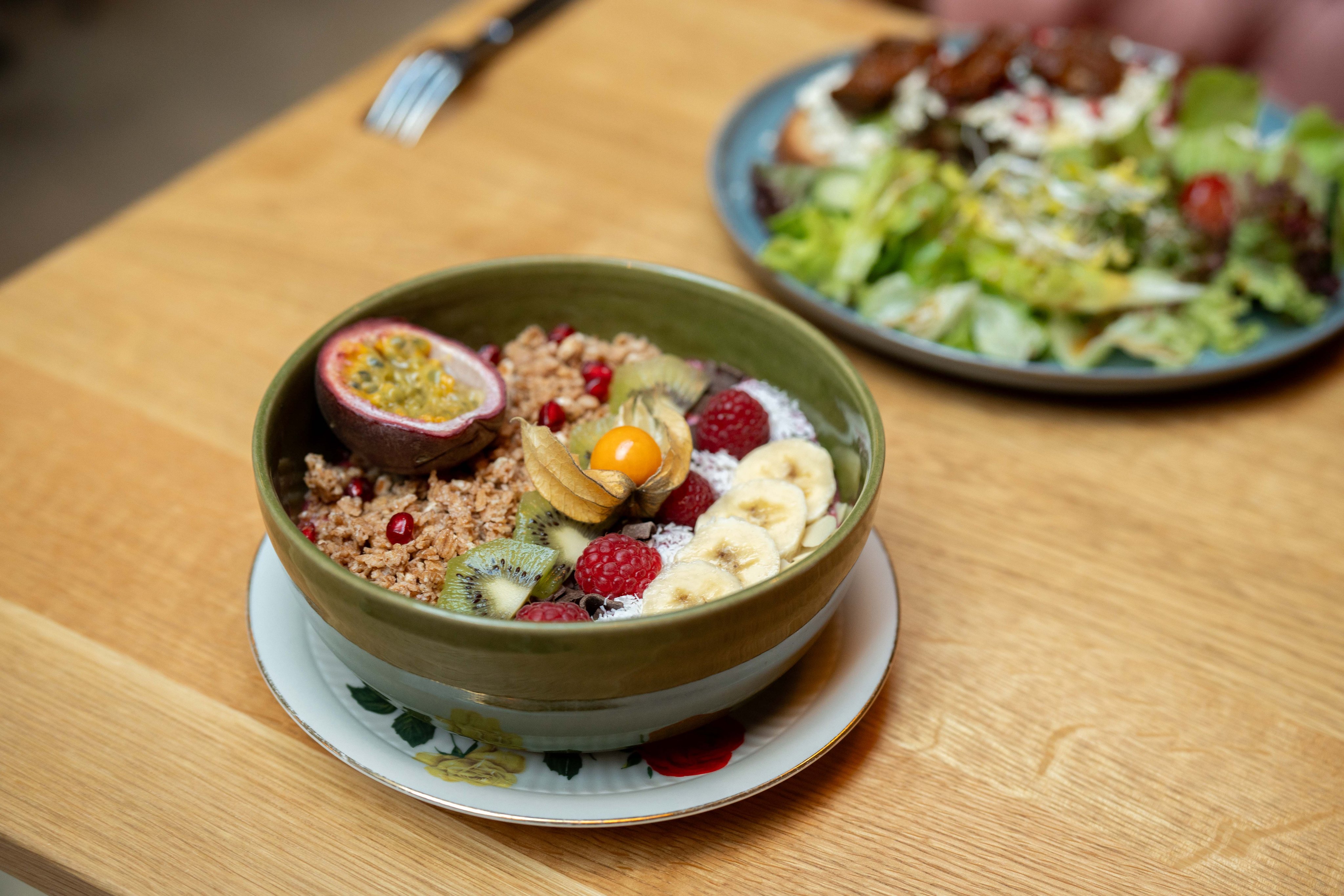 Ein grüner Teller mit Müsli, Obst und Nüssen auf einem Holztisch mit einem Salat im Hintergrund.