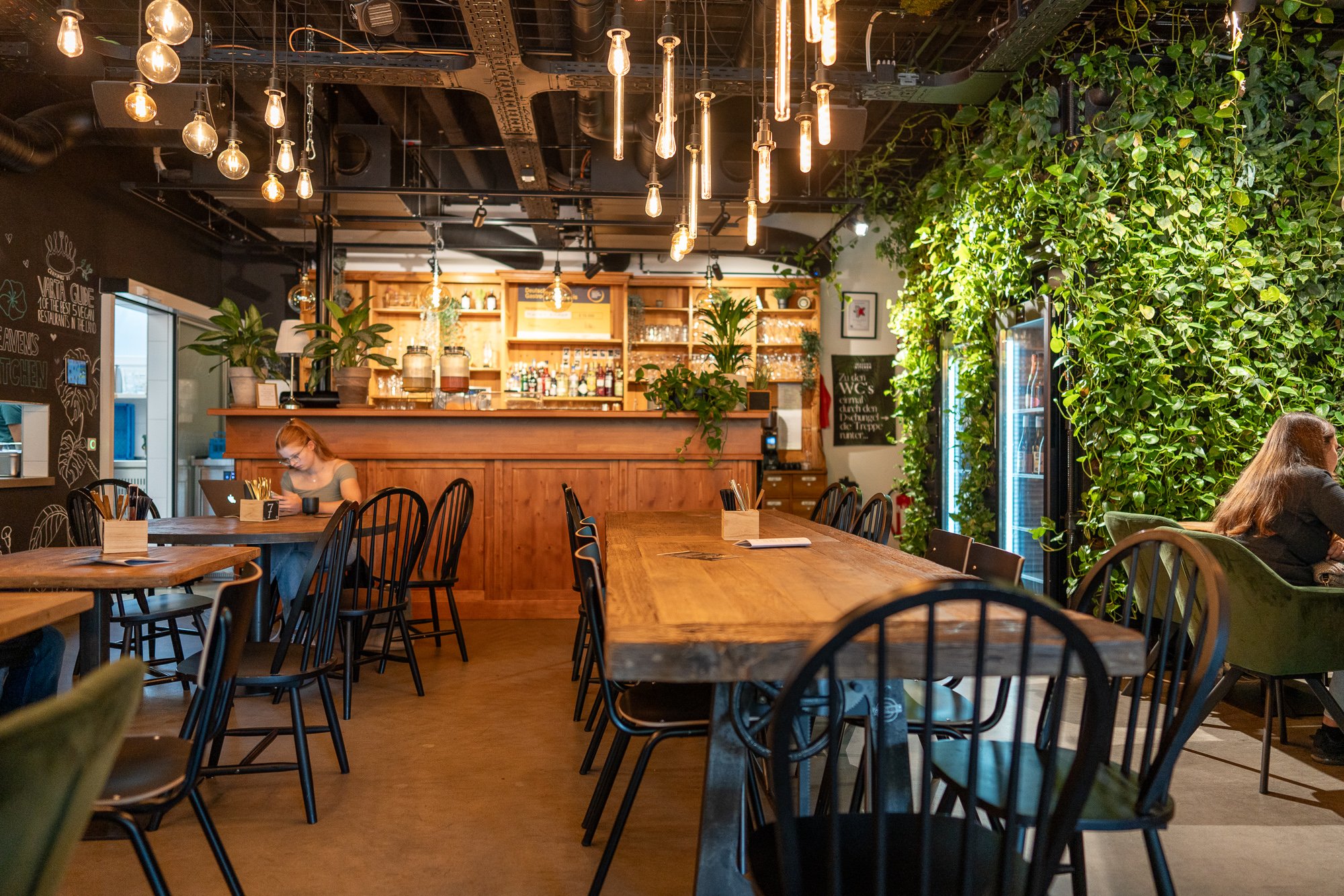 Eine moderne Bar mit einer Holztheke und einer Vielzahl von Flaschen und Gläsern darauf. Die Bar hat verschiedene Sitzmöglichkeiten, darunter Holzstühle und grüne Samtsofas. Die Wände sind mit Tafelkunst und Pflanzen geschmückt. Die Beleuchtung ist industriell und hängt von der Decke. Der Boden ist aus Beton.