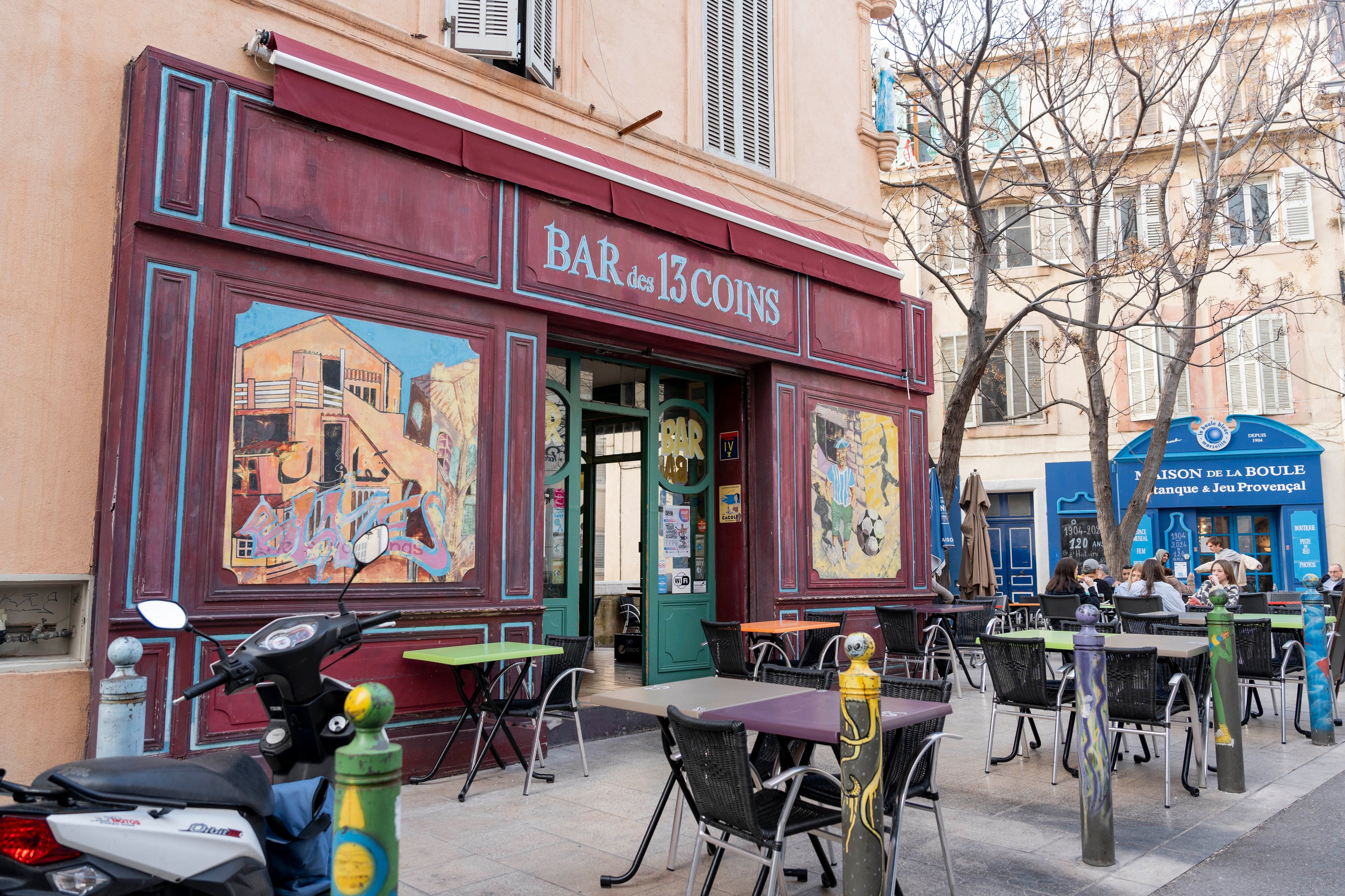 Die Außenansicht der Bar des 13 Coins in Marseille mit bunten Wandmalereien, umgeben von Außensitzplätzen und Bäumen, an einem sonnigen Tag.