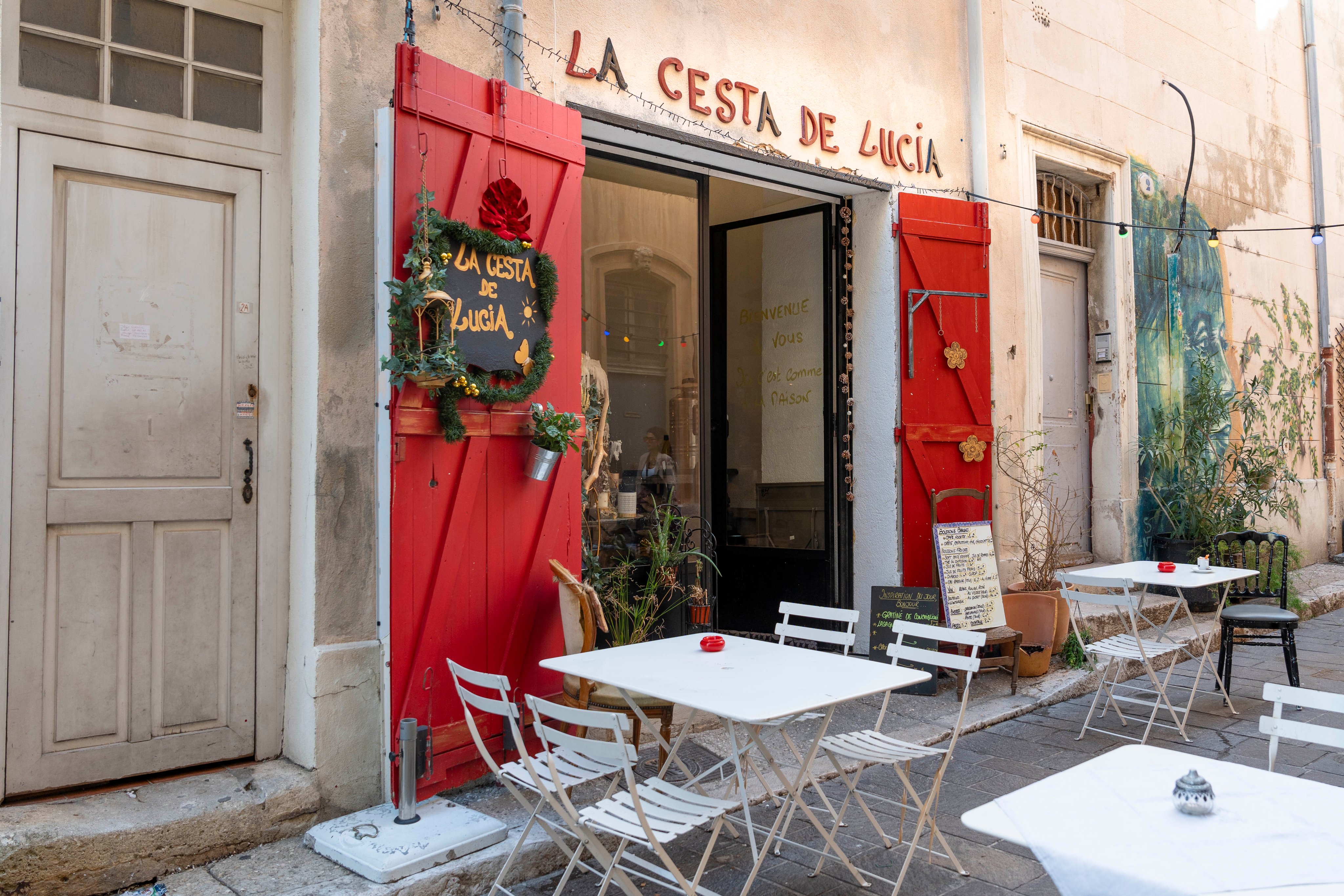 Das Restaurant 'La Cesta de Lucía' in einer ruhigen Straße von Le Panier, Marseille, mit Außensitzgelegenheiten, festlicher Türdekoration und einer Reihe bunter Lichter.