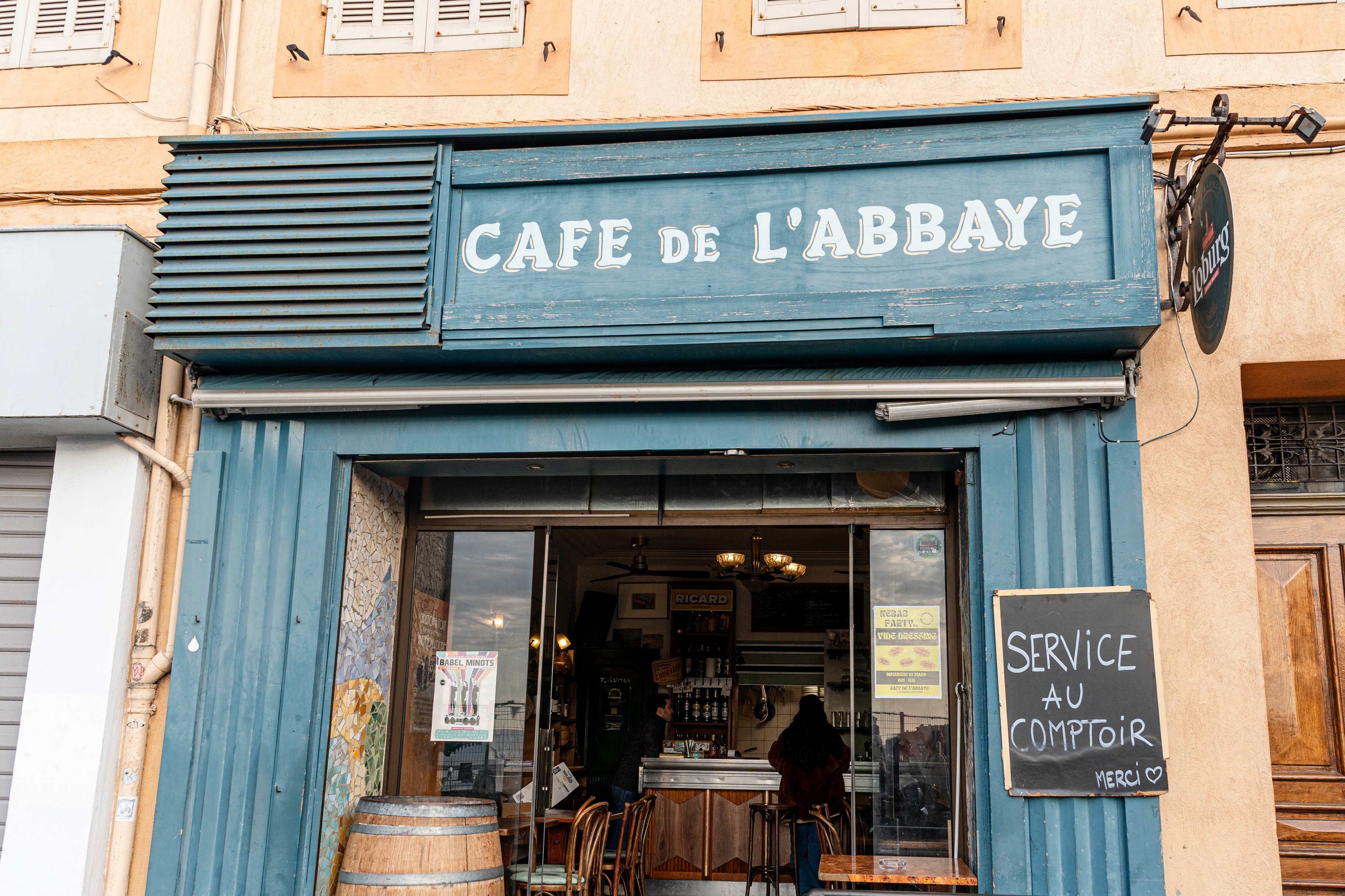 Vorderansicht des Café de l'Abbaye in Marseille, mit traditioneller blauer Markise und Schildern, die freundlich zum Service am Tresen einladen.