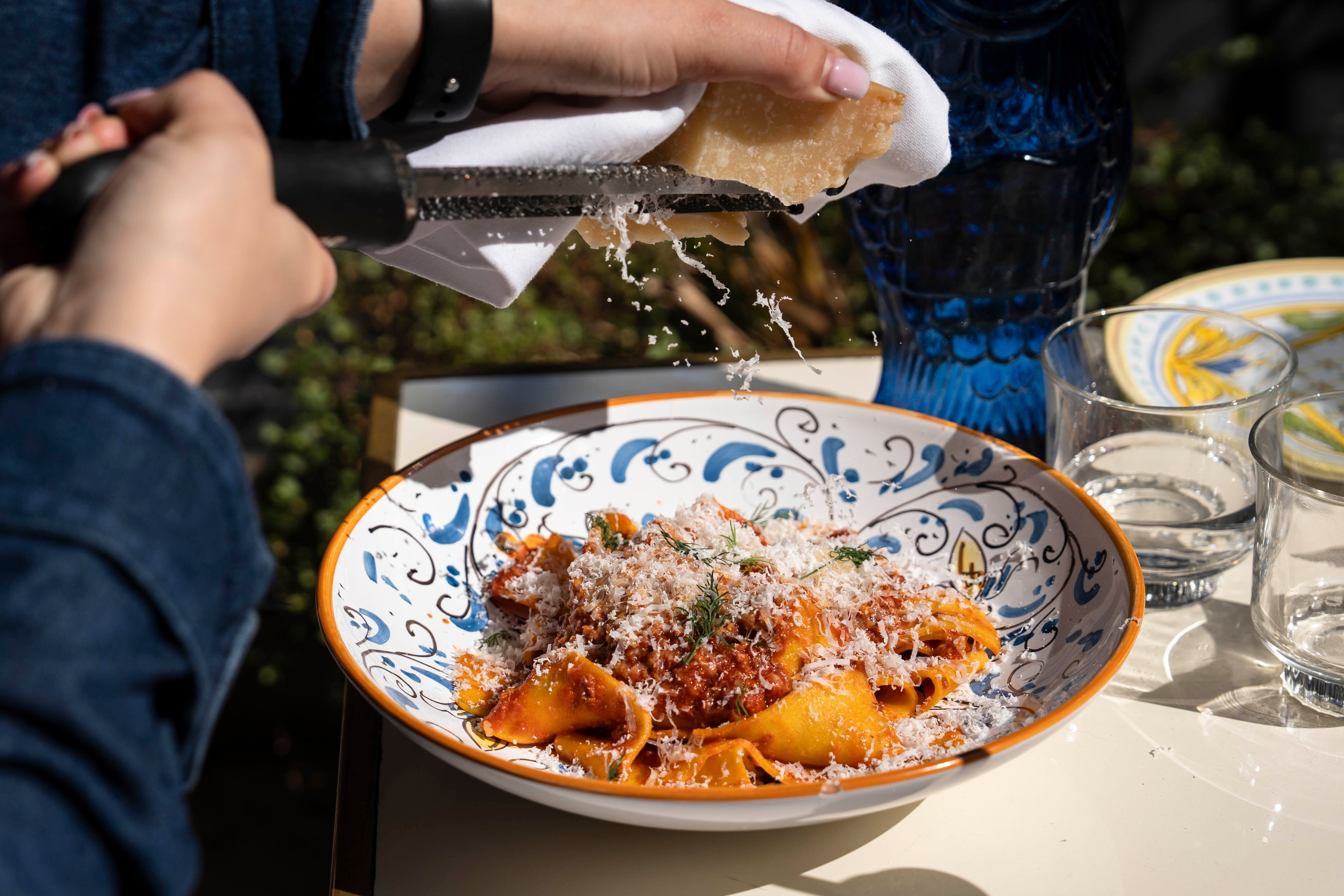 Hände, die über einer Schüssel mit italienischer Pasta Parmesankäse reiben, mit einem Muster des Geschirrs, das den Charme der Mahlzeit hervorhebt.