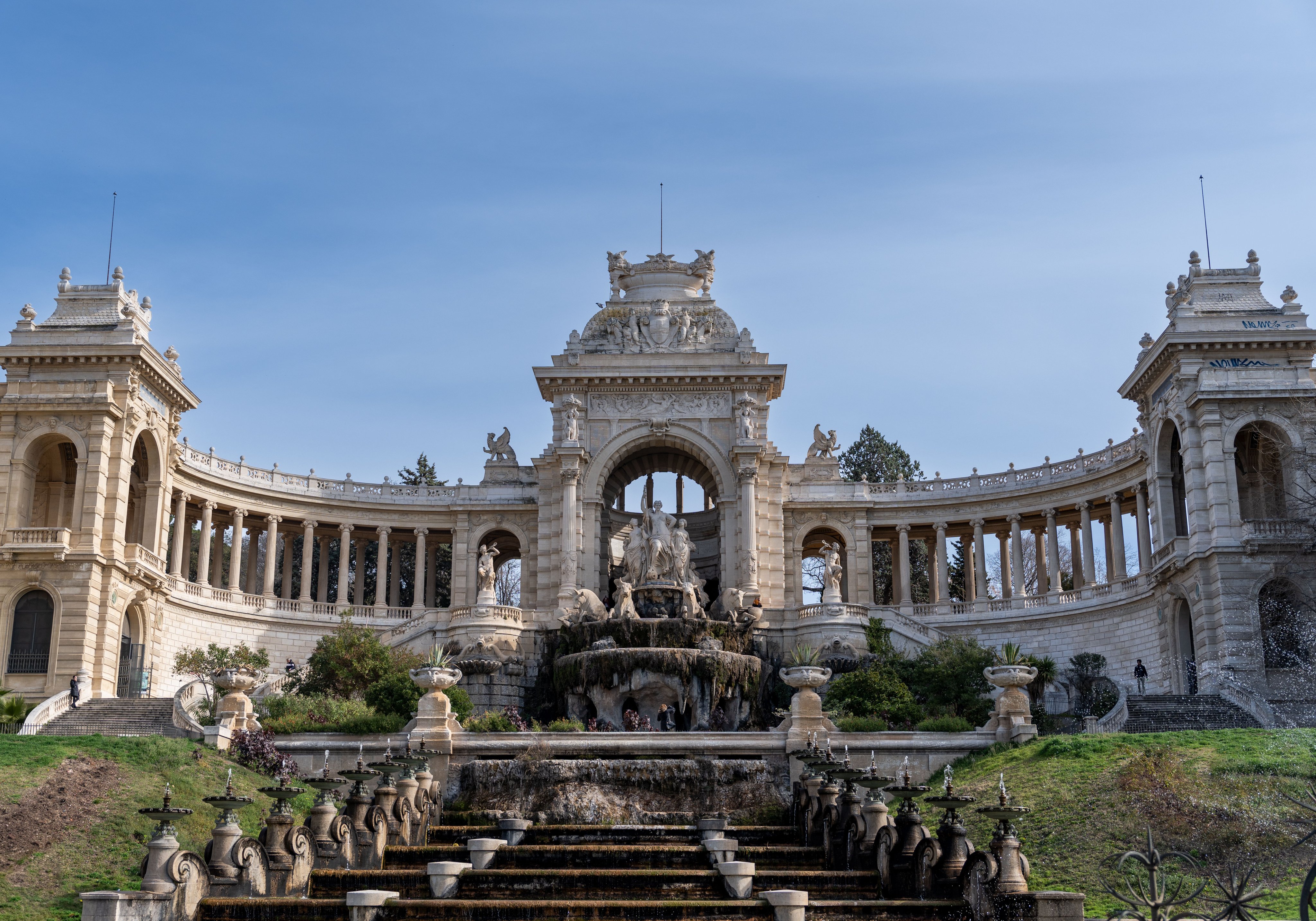 Ansicht des majestätischen Palais Longchamp in Marseille, mit seiner beeindruckenden Fontäne, umgeben von eleganten Säulengängen und Skulpturen unter klarem blauem Himmel.