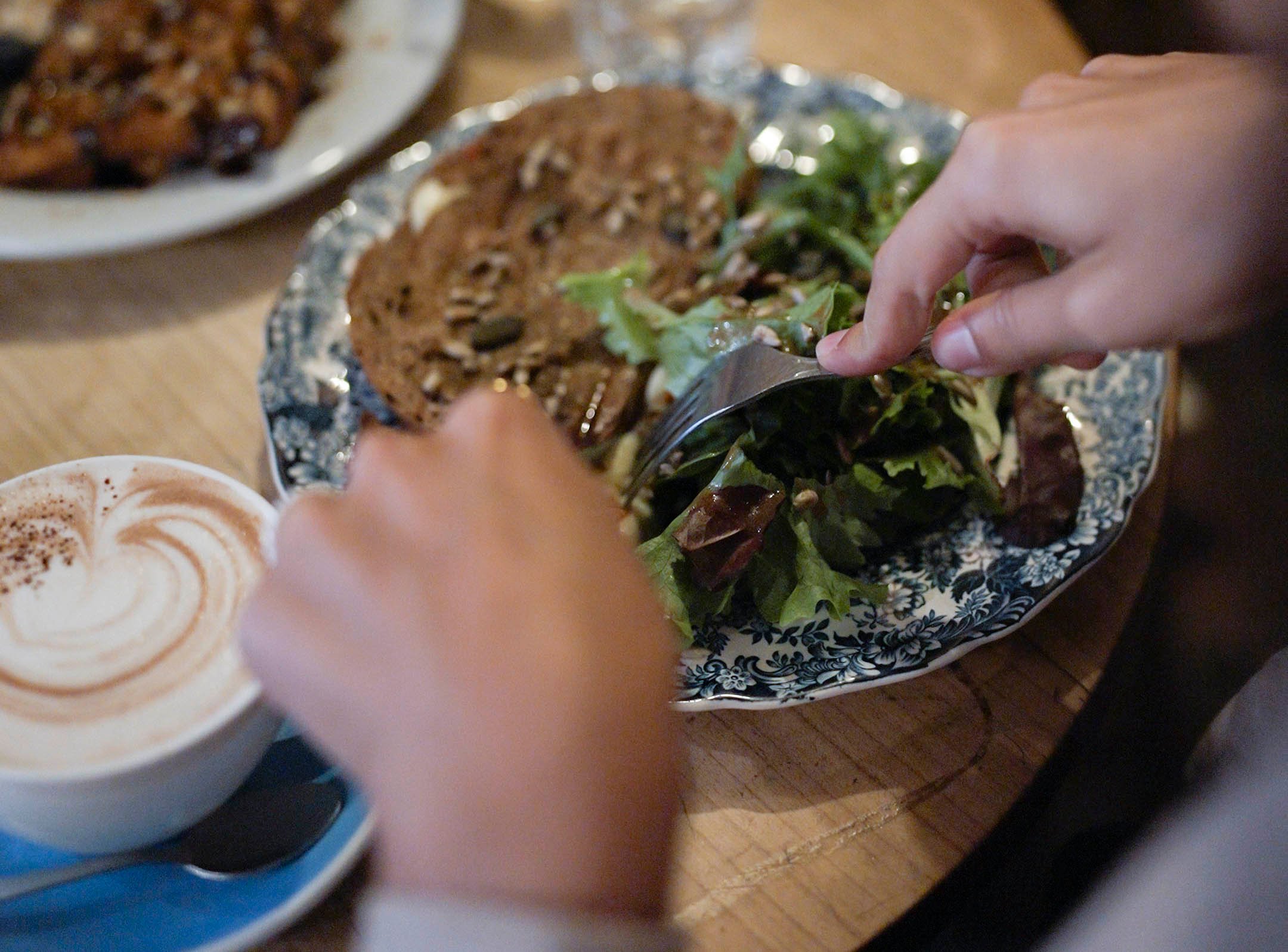 Eine Hand, die einen Salat neben einem kunstvoll angerichteten Toast mit Käse undgetrockneten Tomaten zubereitet, neben einer Tasse Cappuccino mit Herz-Schaumkrone.