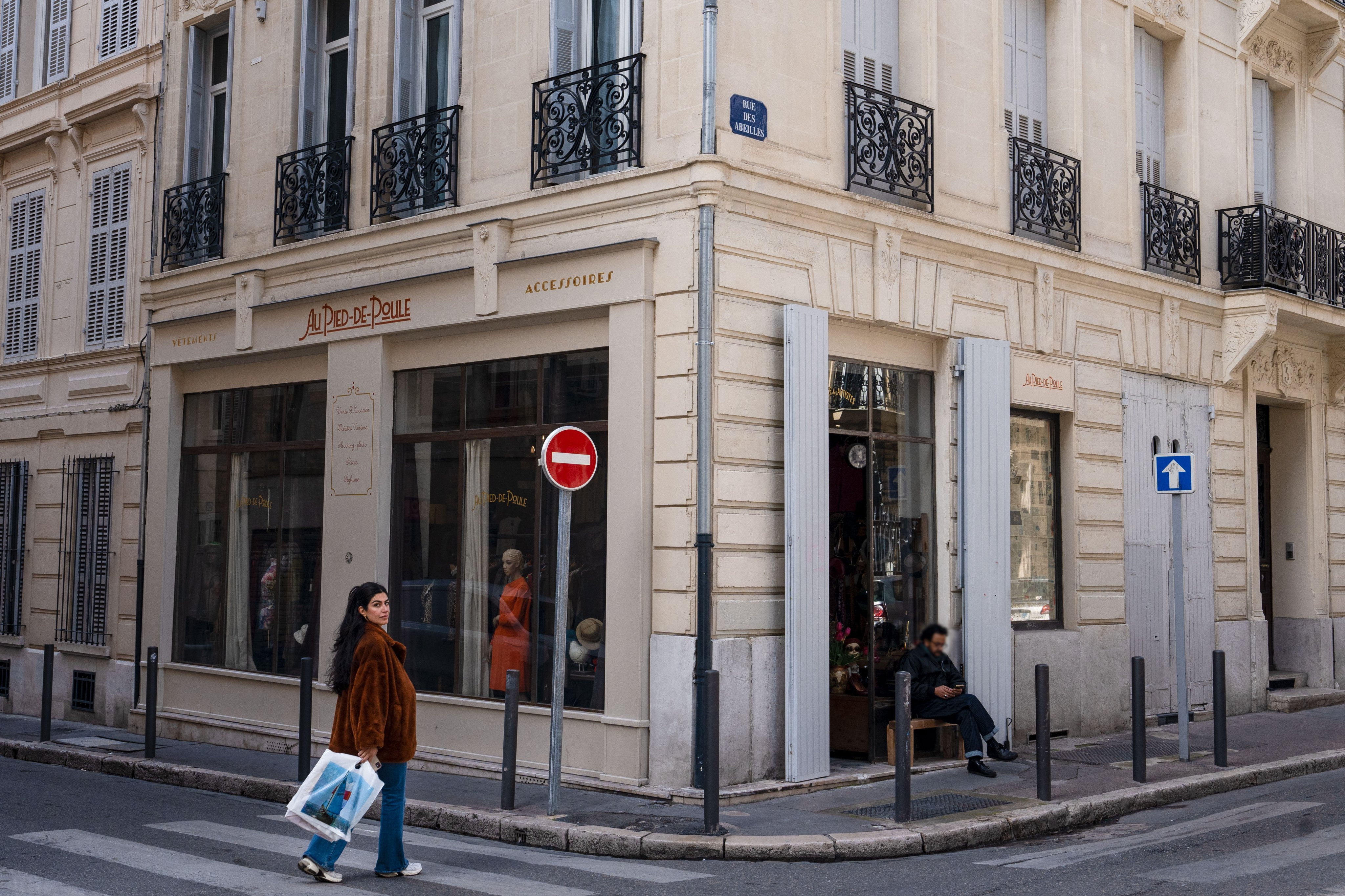 Eine Passantin geht an dem Eckladen 'Au Pied de Poule' vorbei, einem Geschäft für Secondhand-Mode in Marseille, mit einer Vielzahl von Kleidungsstücken sichtbar durch das Schaufenster.