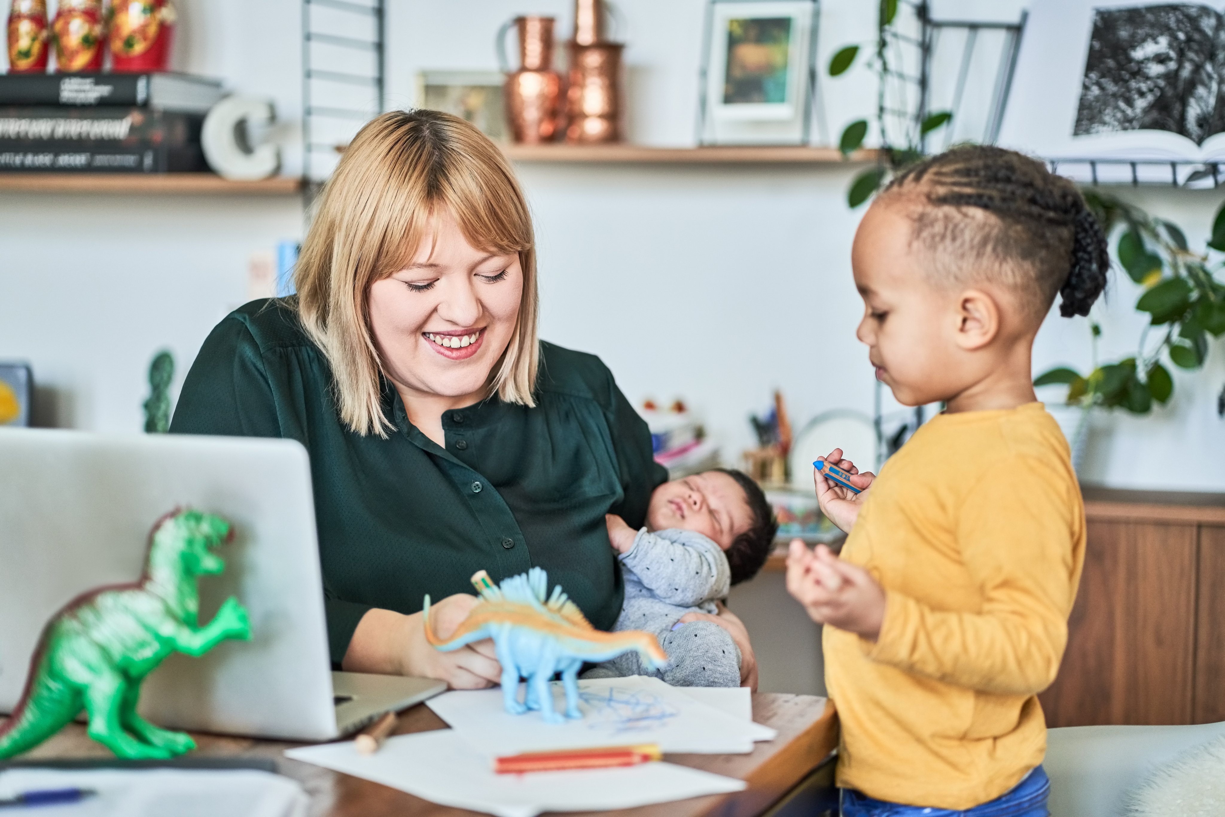 Smiling mother holds newborn, plays with son while working from home.