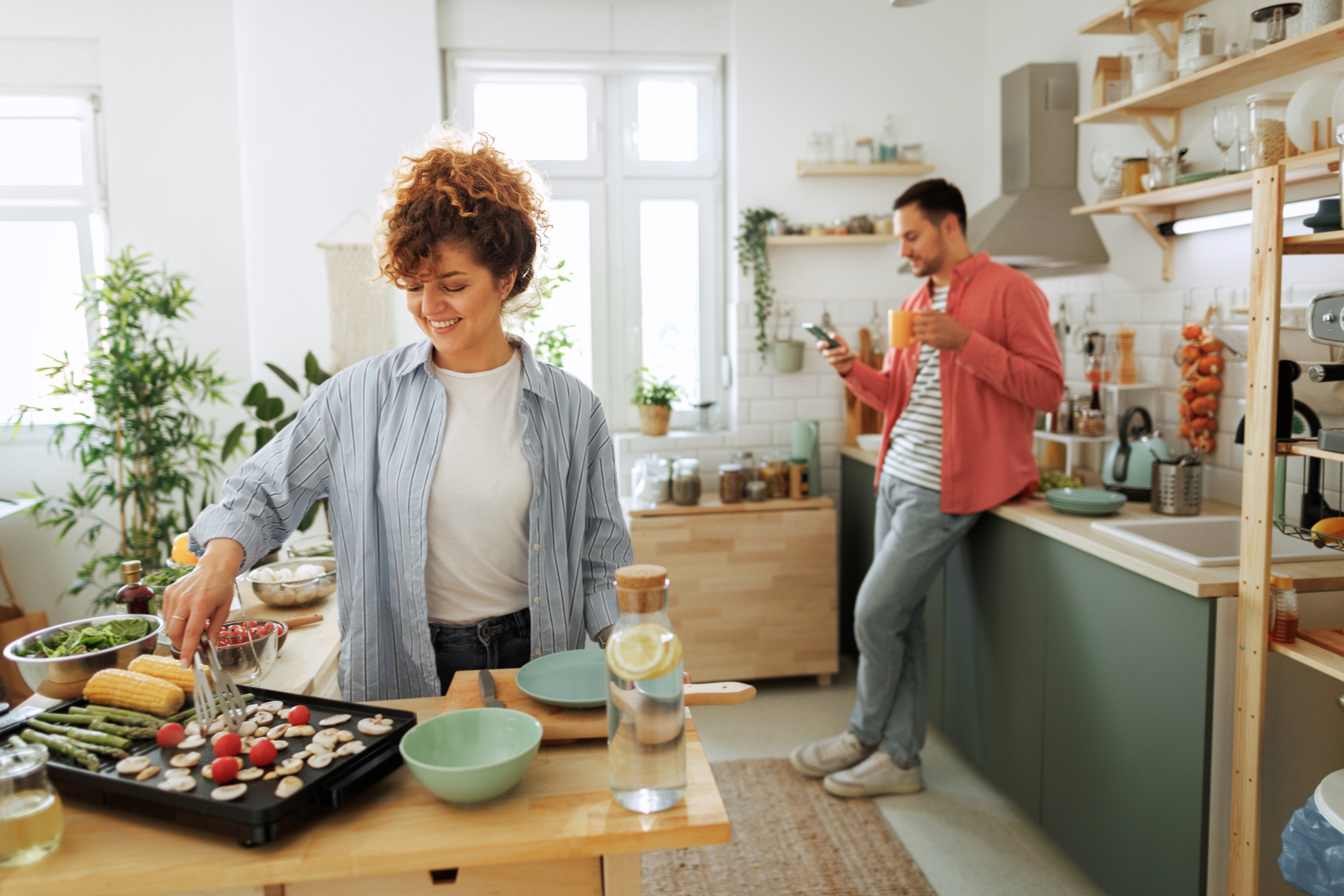 A young woman cooks in the kitchen while a man looks at his phone.