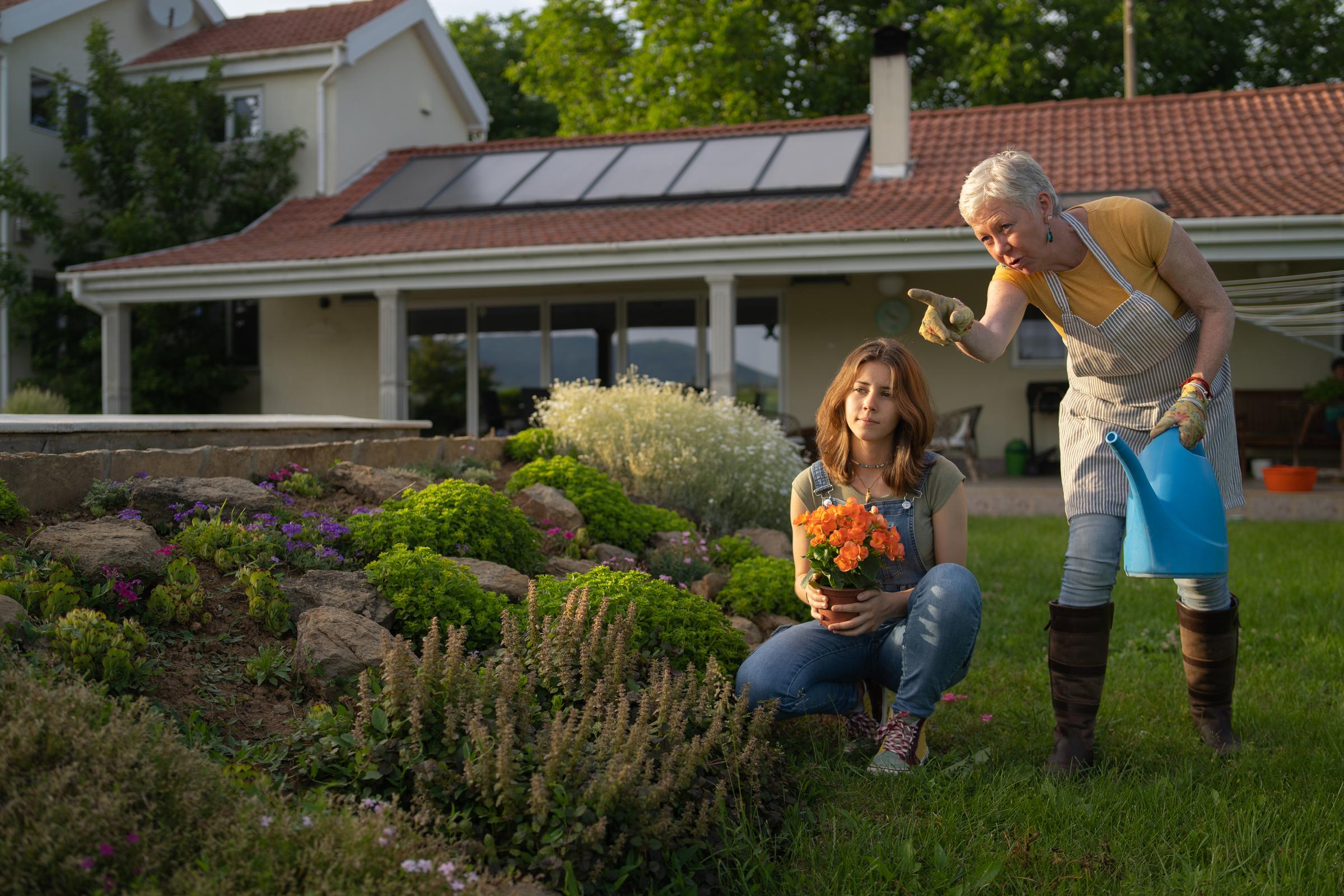 Grandmother points while granddaughter looks on, while holding flower pot in a garden