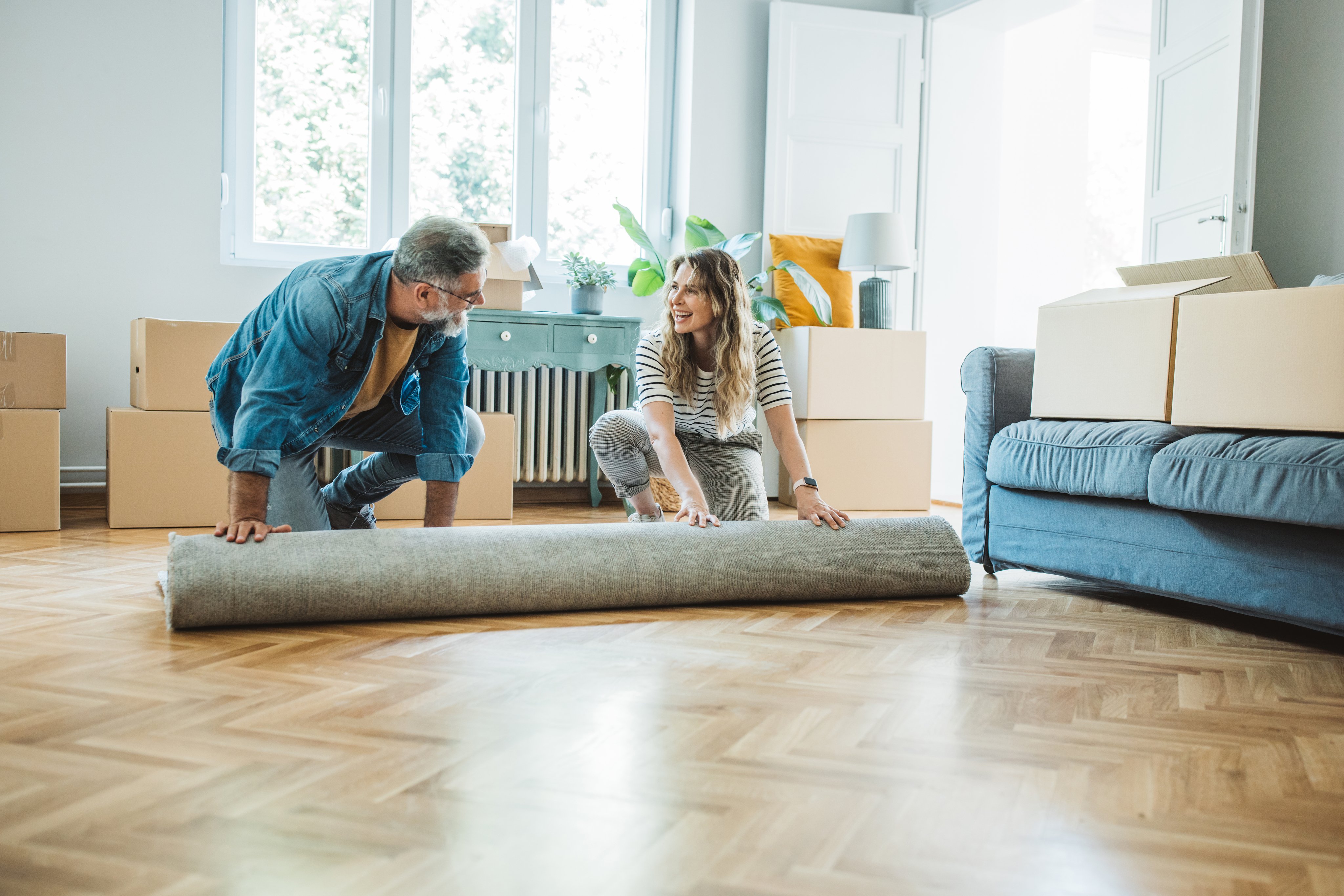 A young woman and her father unroll a rug. They're surrounded by packing boxes.