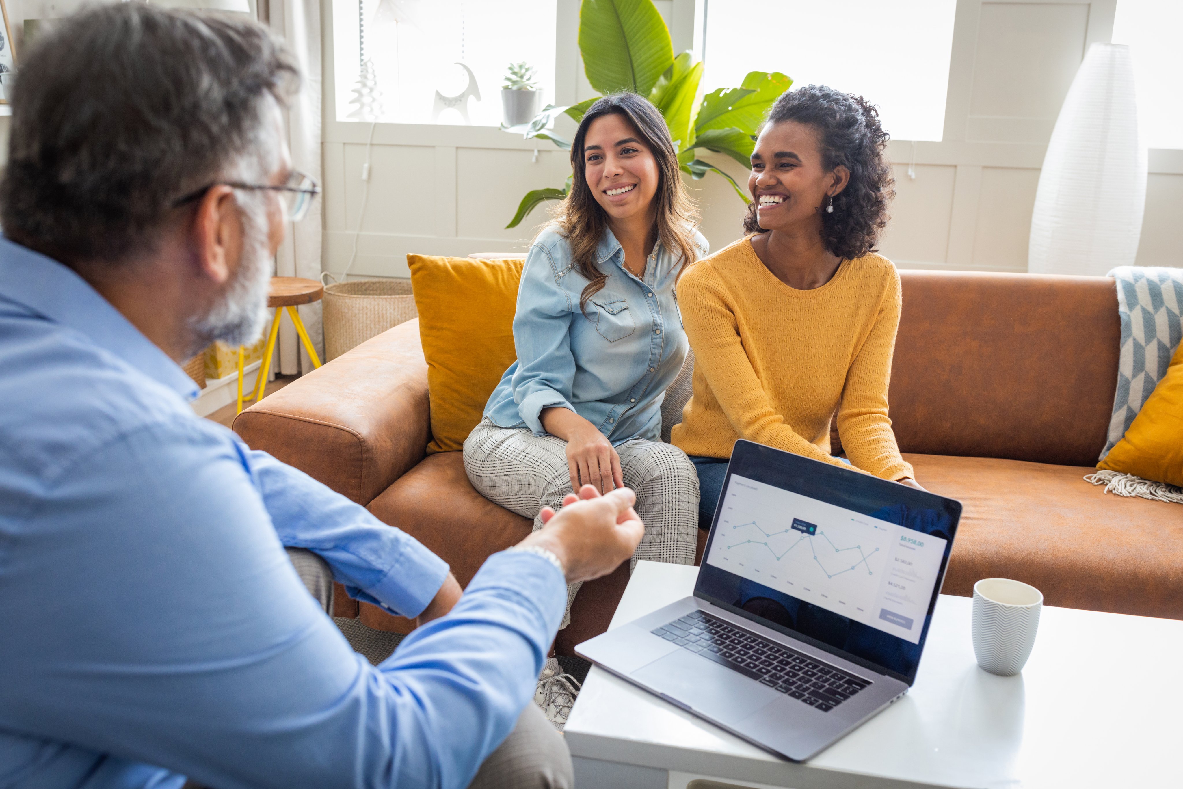 Two young women sit smiling opposite a mortgage broker. His laptop shows some figures and graphs.