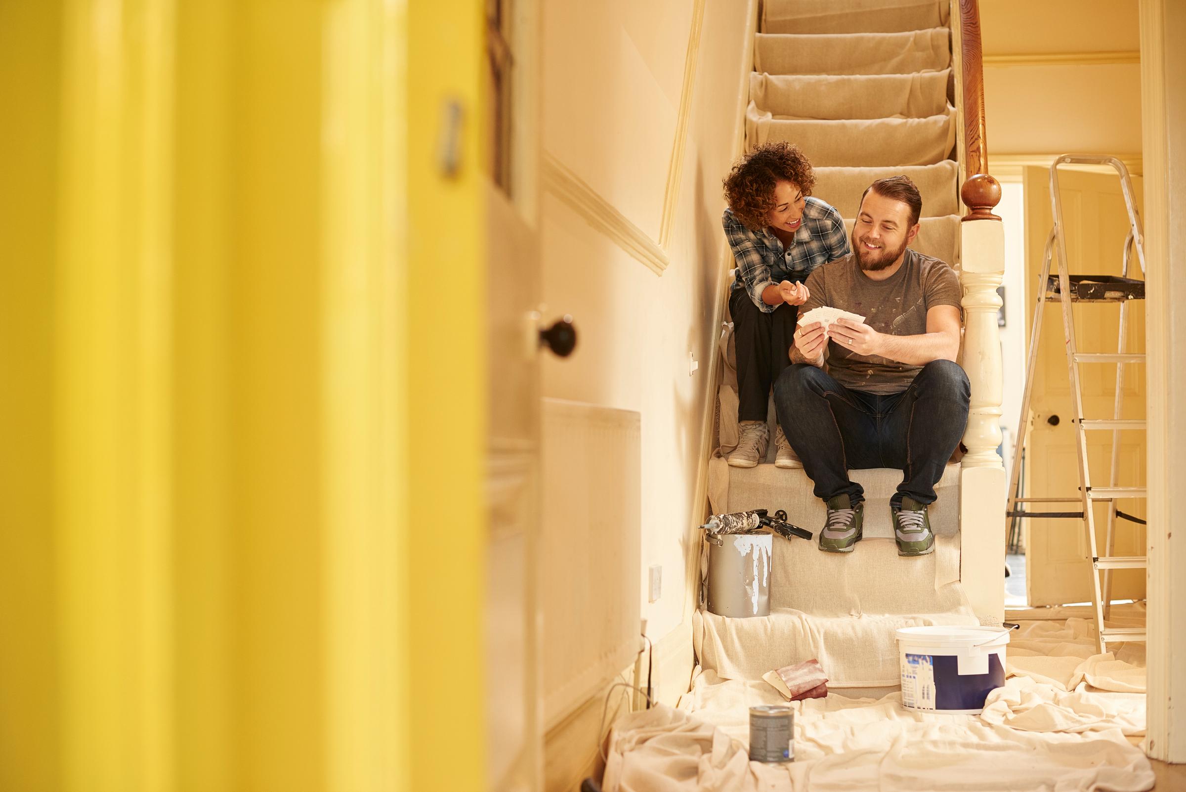 A couple sits on their stairs looking at paint colour cards, surrounded by decorating tools.