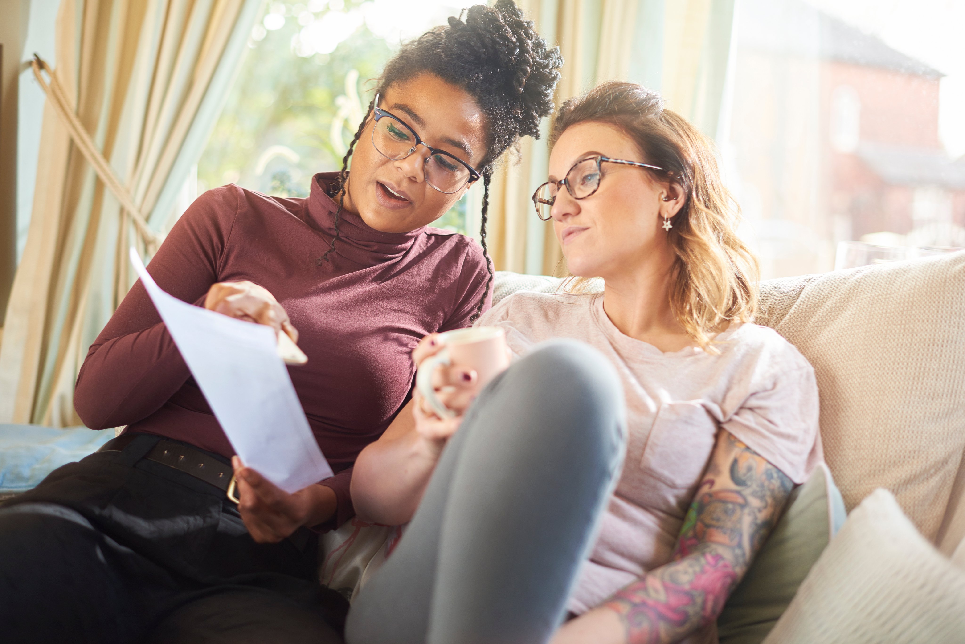 Two females are sat on a sofa. One female holds a mug and the other the piece of paper.  