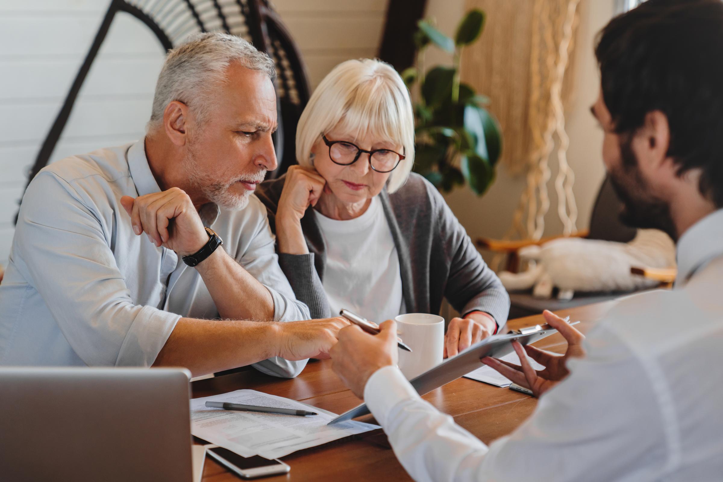 A mature couple is sitting at a table. A businessman is showing them a touchpad screen.