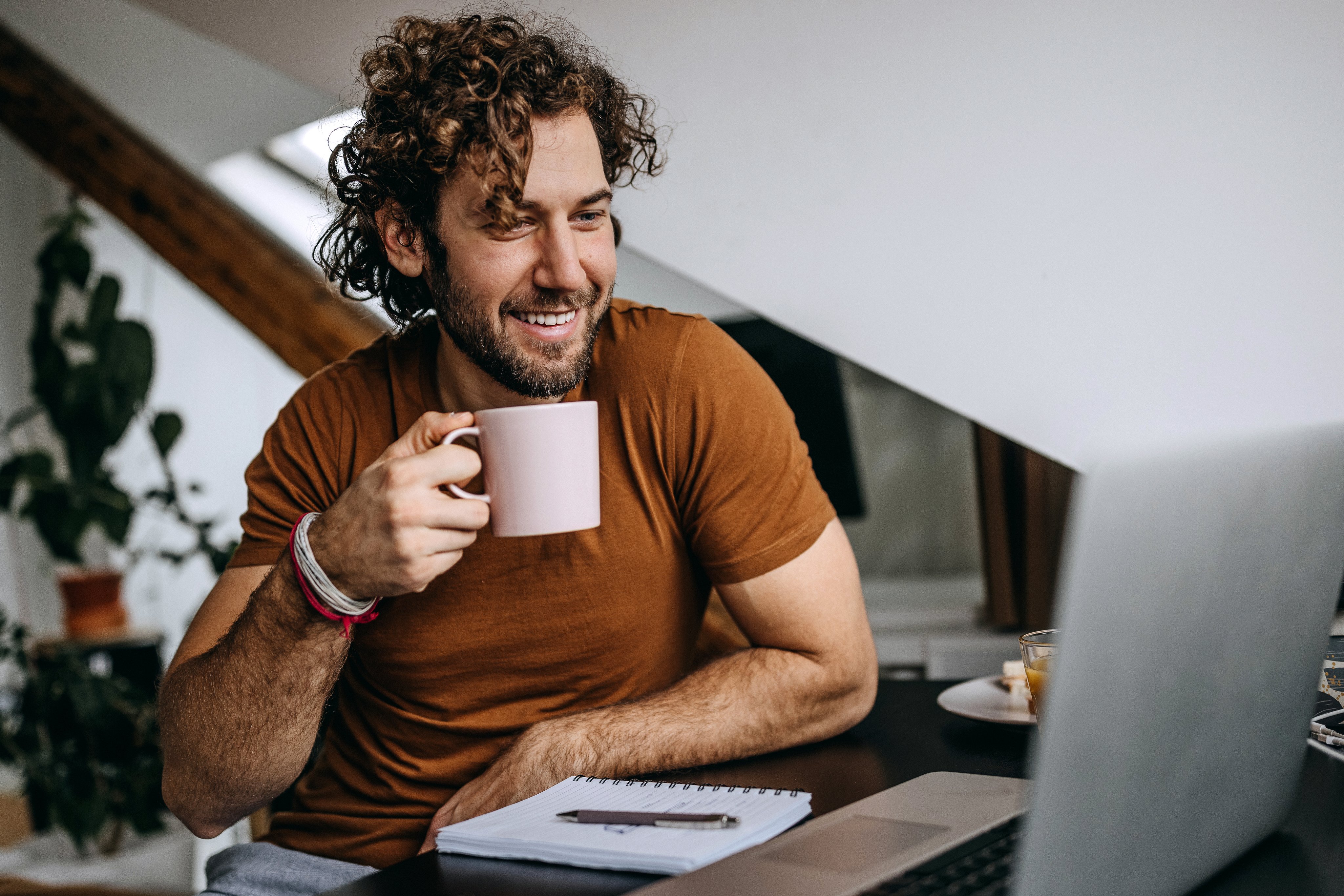 A male is sat at a desk drinking coffee and looking at his laptop.