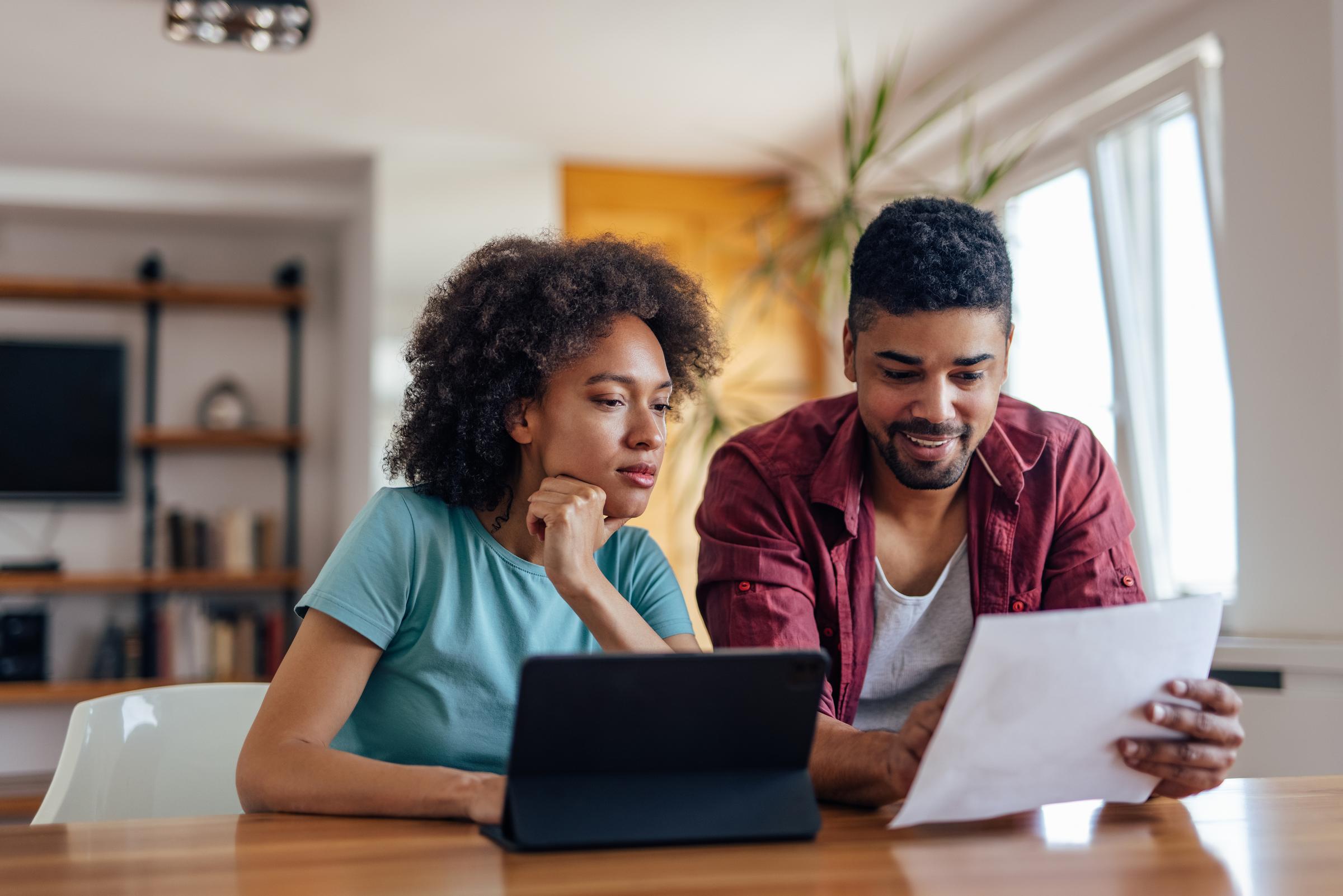  A man and a woman are sat at a dining table. They are looking at a piece of paper and a touchpad. 