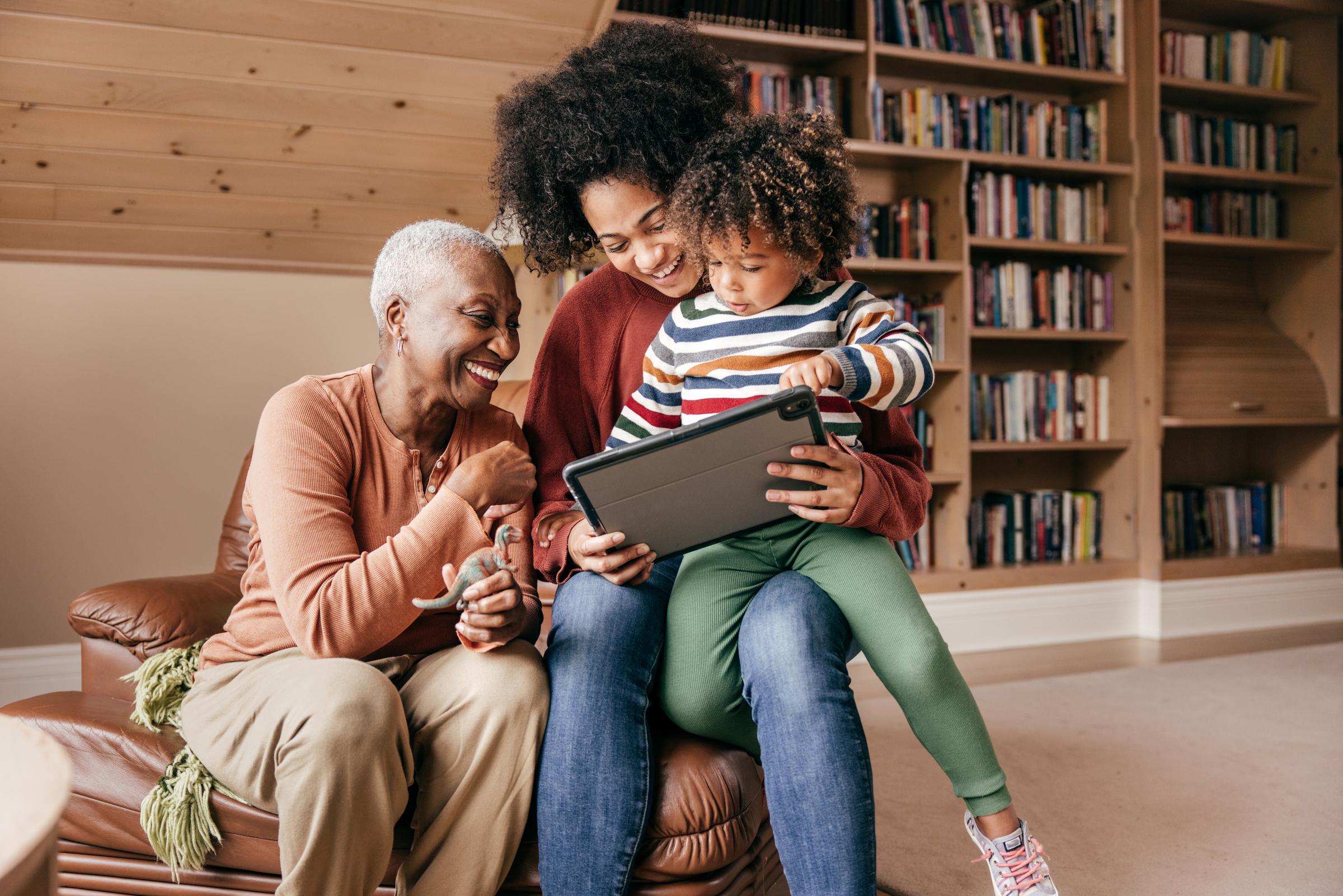 A grandmother, mother and child sit on an armchair and smile at a tablet screen. 