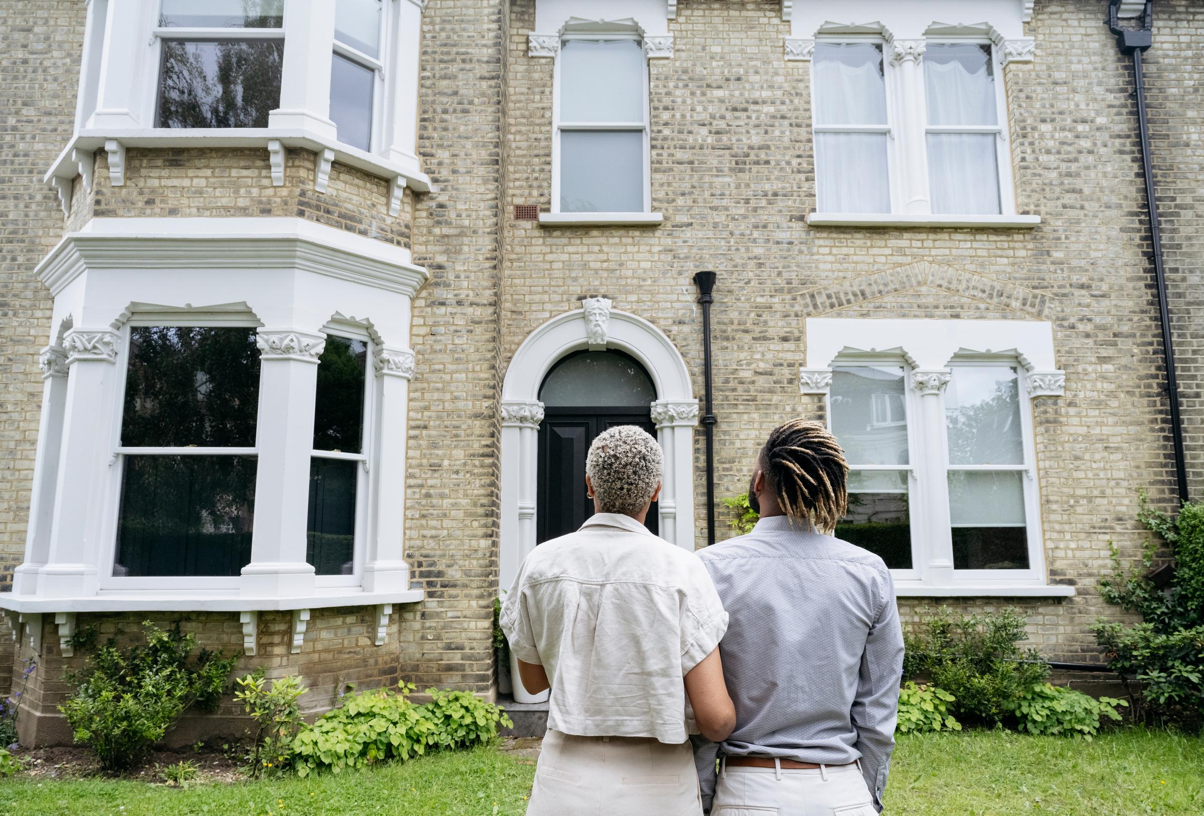 A young couple is looking at a large Victorian home. They are linking arms. 