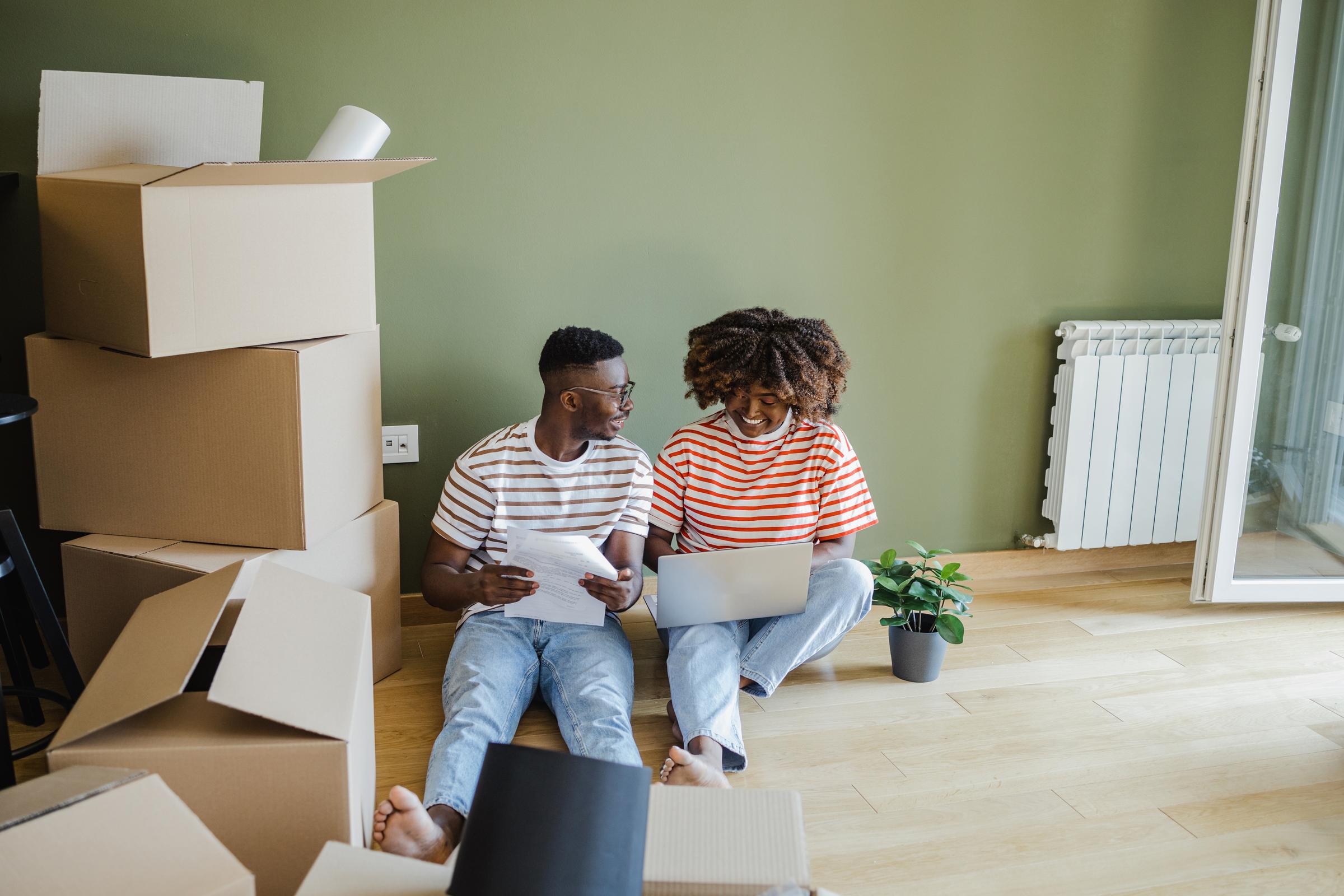 Couple sit on the floor with removal boxes. Woman holds laptop and man holds a piece of paper.