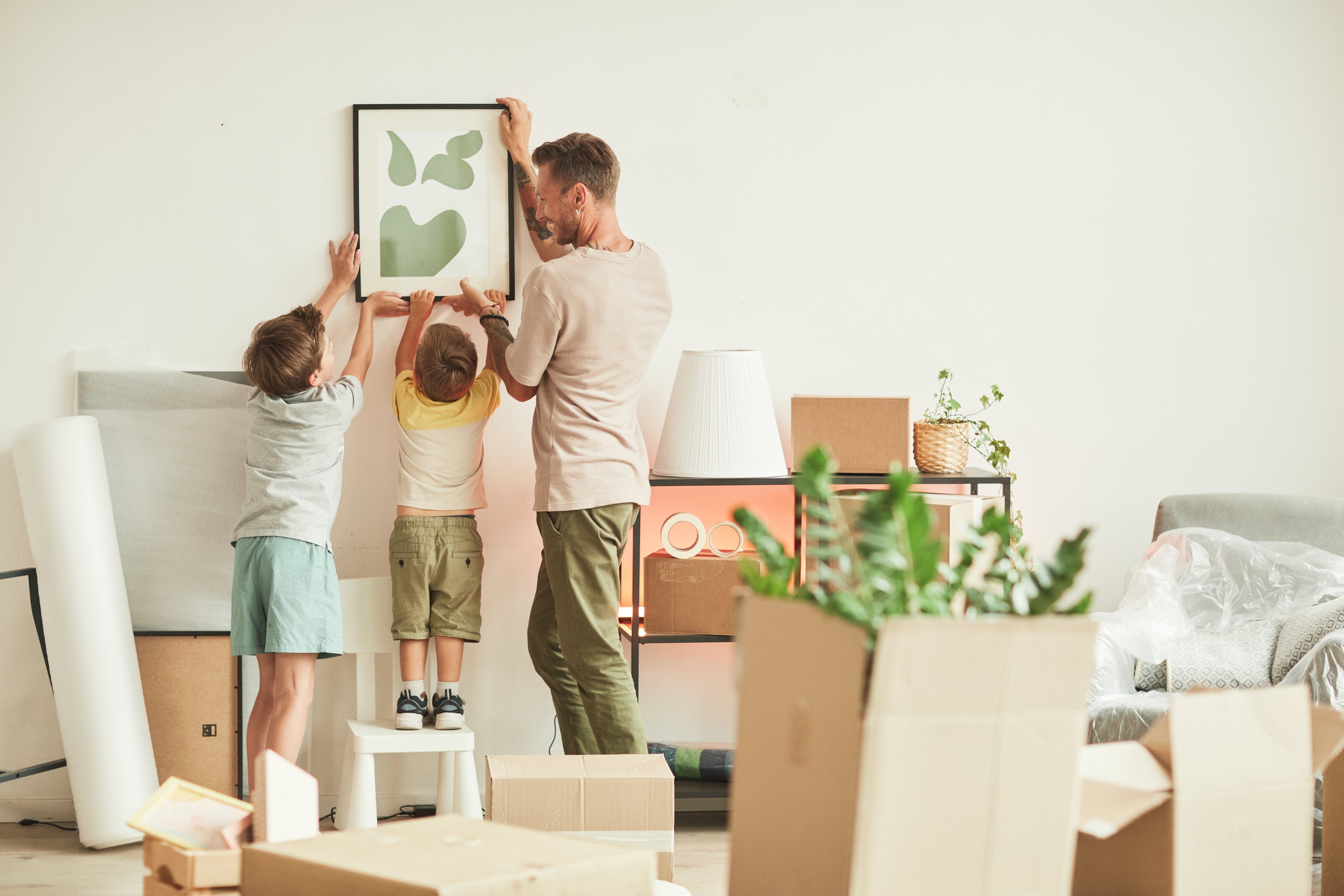 Dad and his 2 children are deciding where to hang a picture. They have moving boxes around the room.