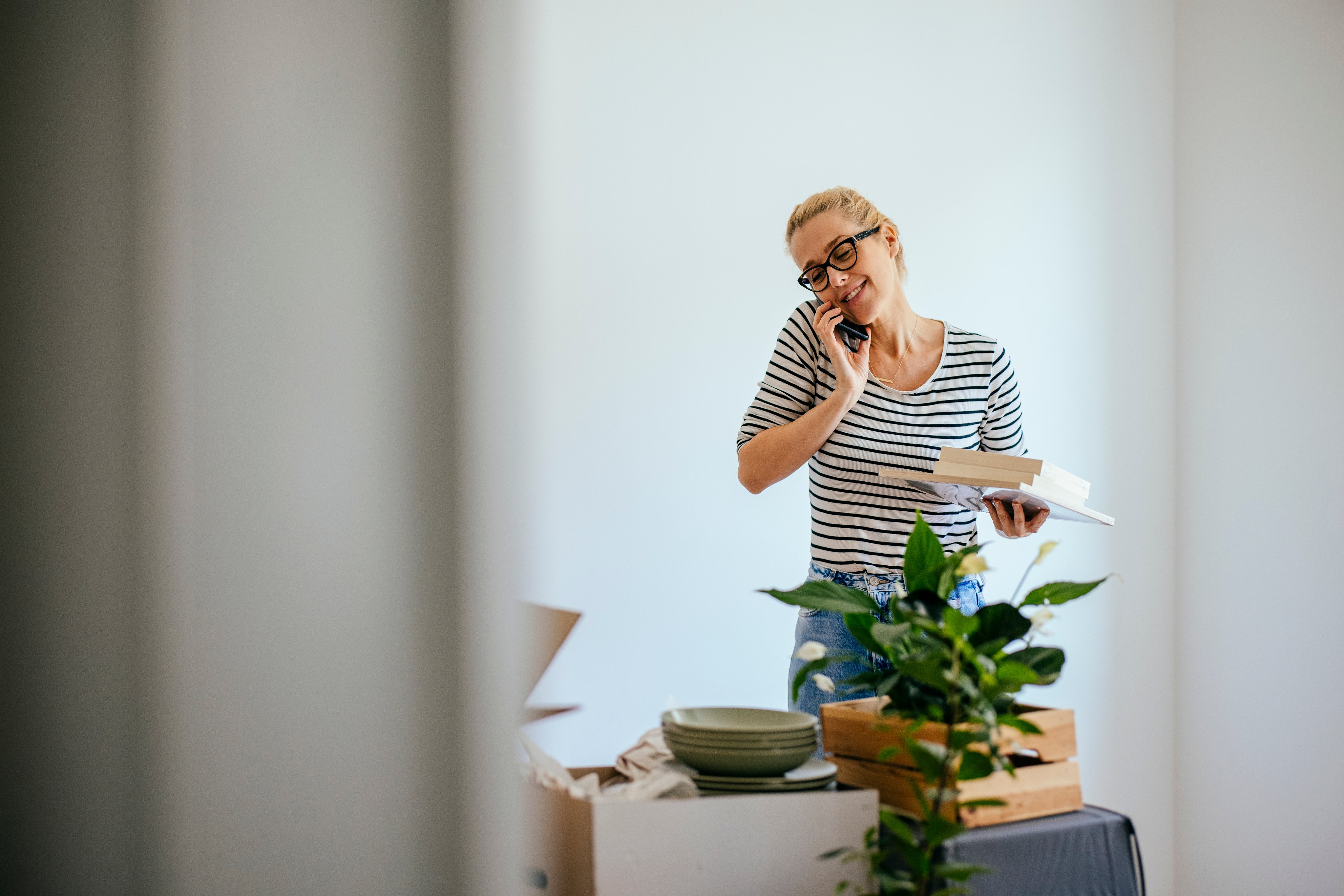 A woman with glasses is on the phone while unpacking. Boxes contain plants,  kitchenware and books. 