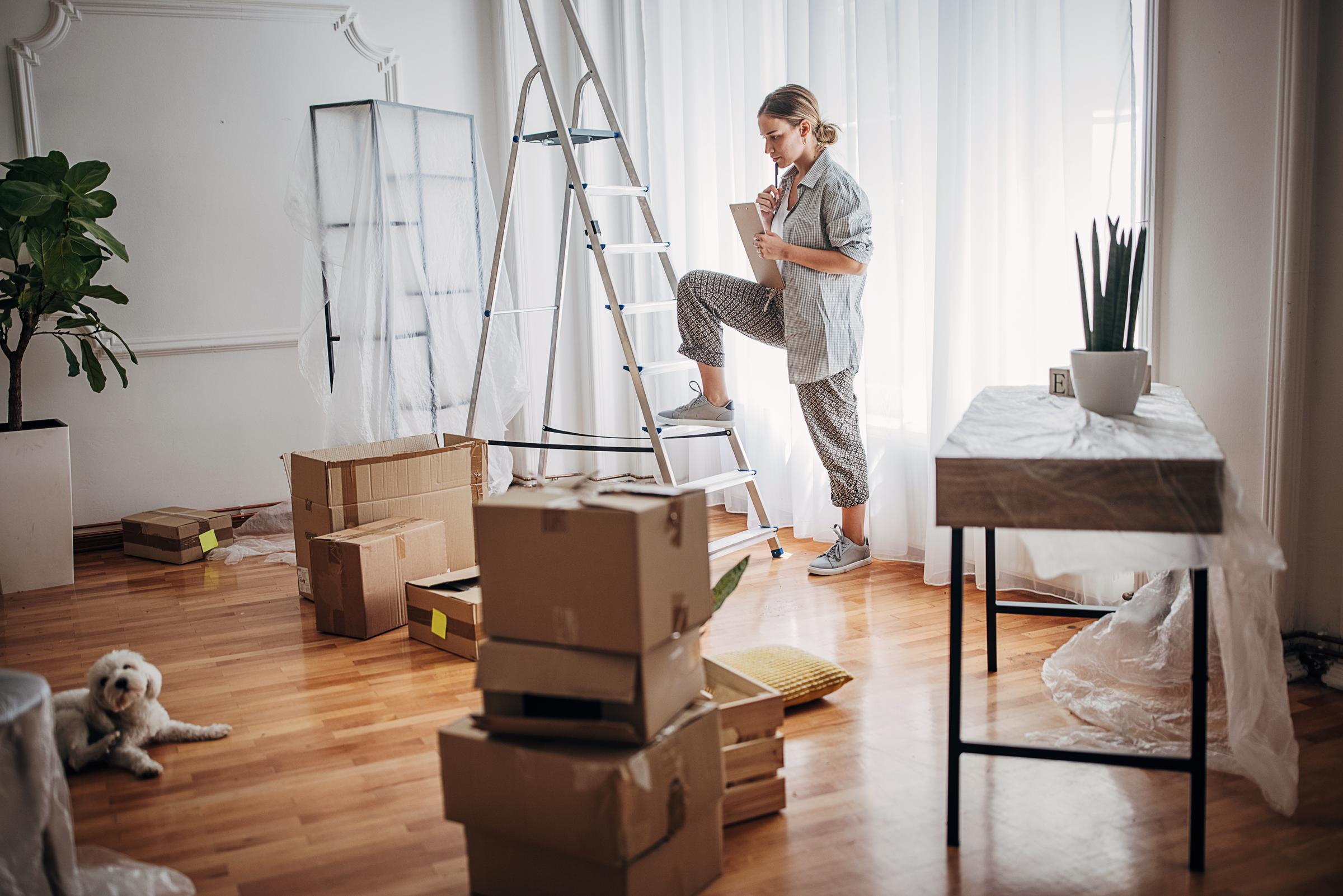 Woman has foot on ladder, thinking, she is decorating. Her white dog relaxes on the floor. 